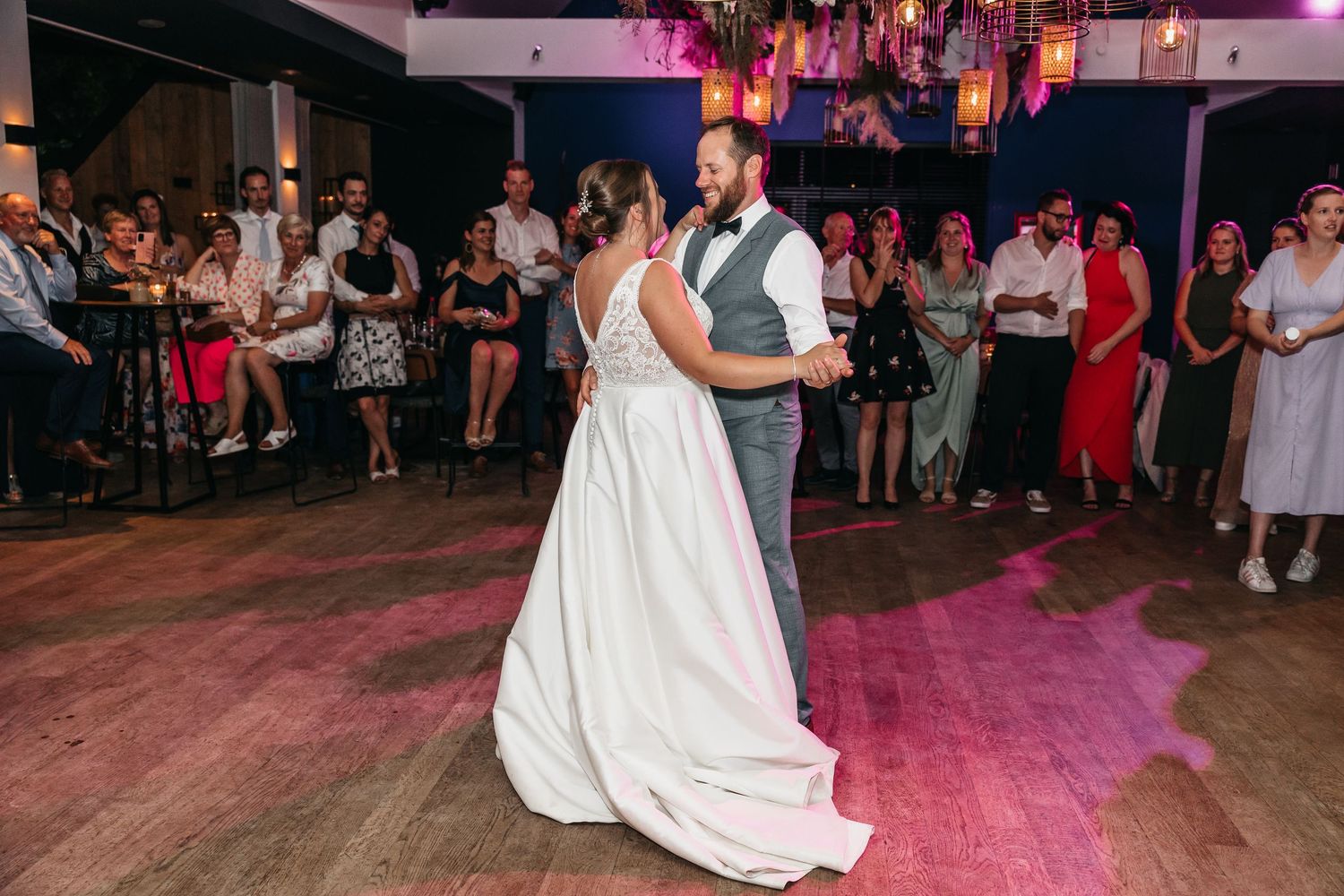 Bride and groom sharing first dance before guests on pink-lit wedding reception dance floor.