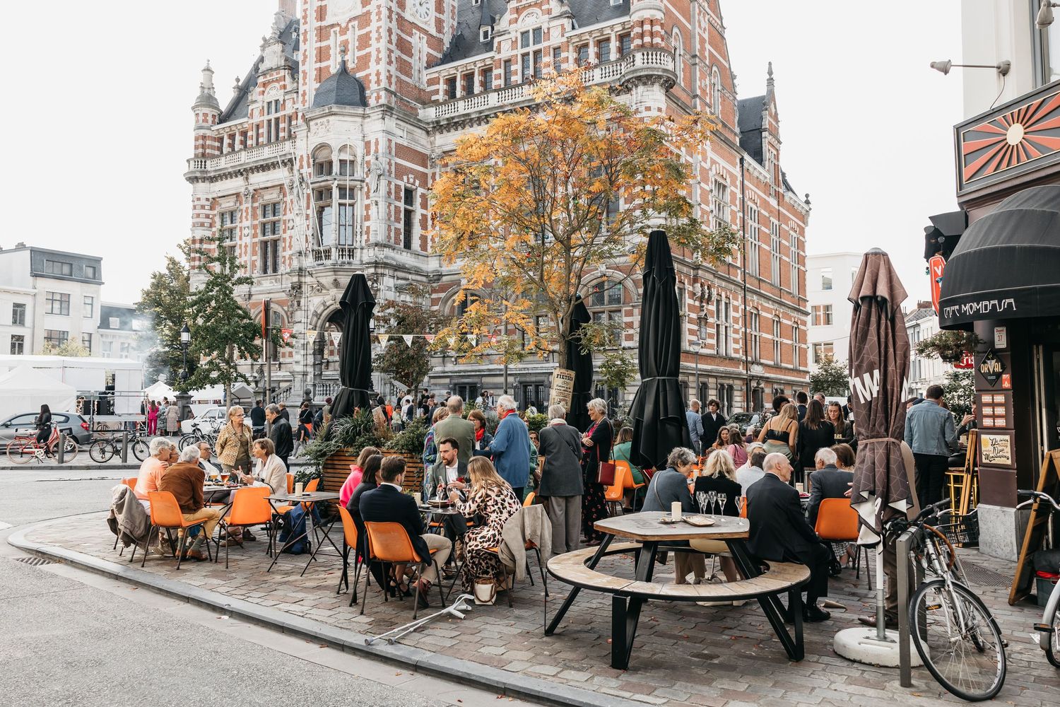 Crowds of people enjoying outdoor terrace seating at a café in a European city square with autumn trees.