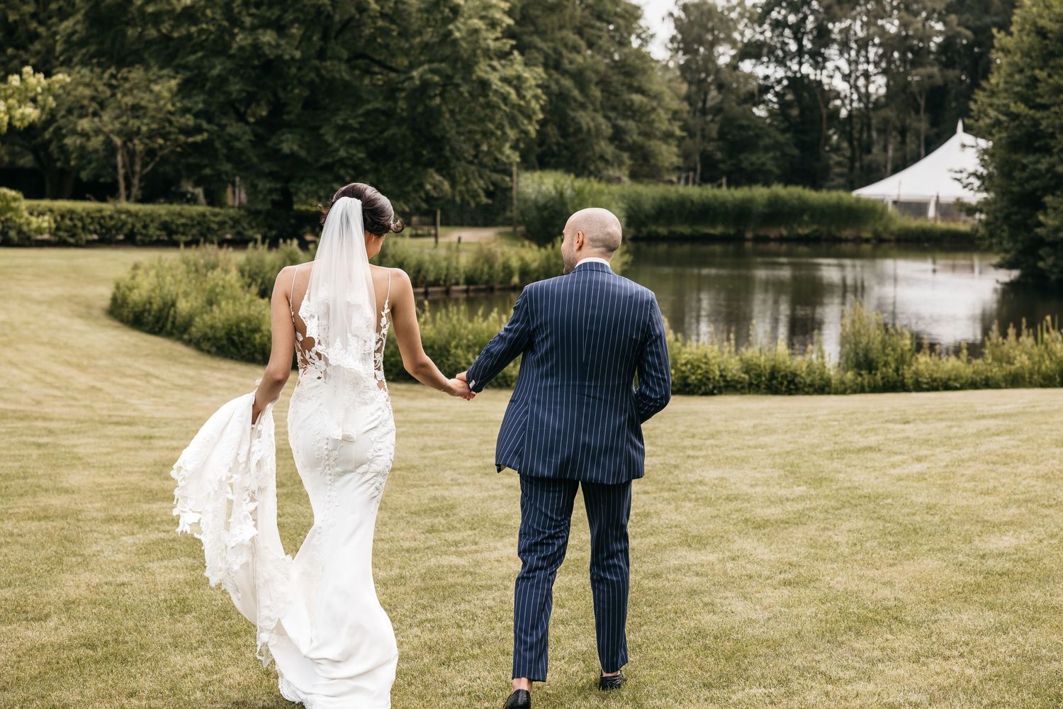 Bride in lace gown and veil walks hand-in-hand with suited guest toward a pond in a lush garden.