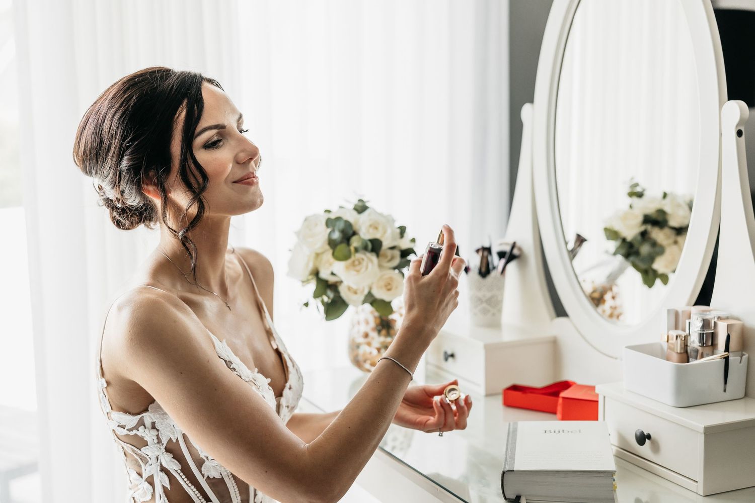 Beautiful bride applying perfume at a white vanity table with white floral bouquet and oval mirror.