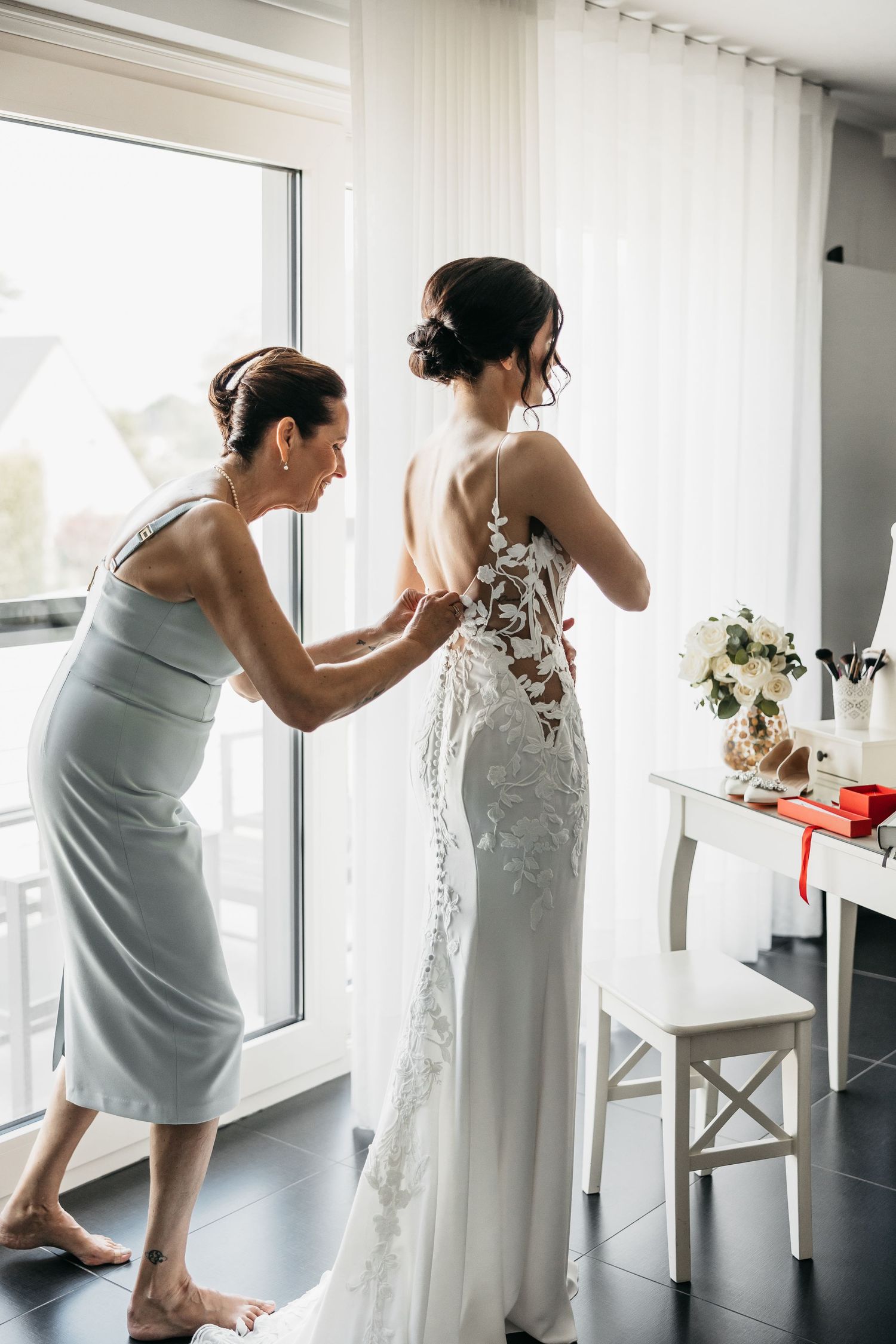 Mother helping bride button up her lace wedding dress near a bright window.