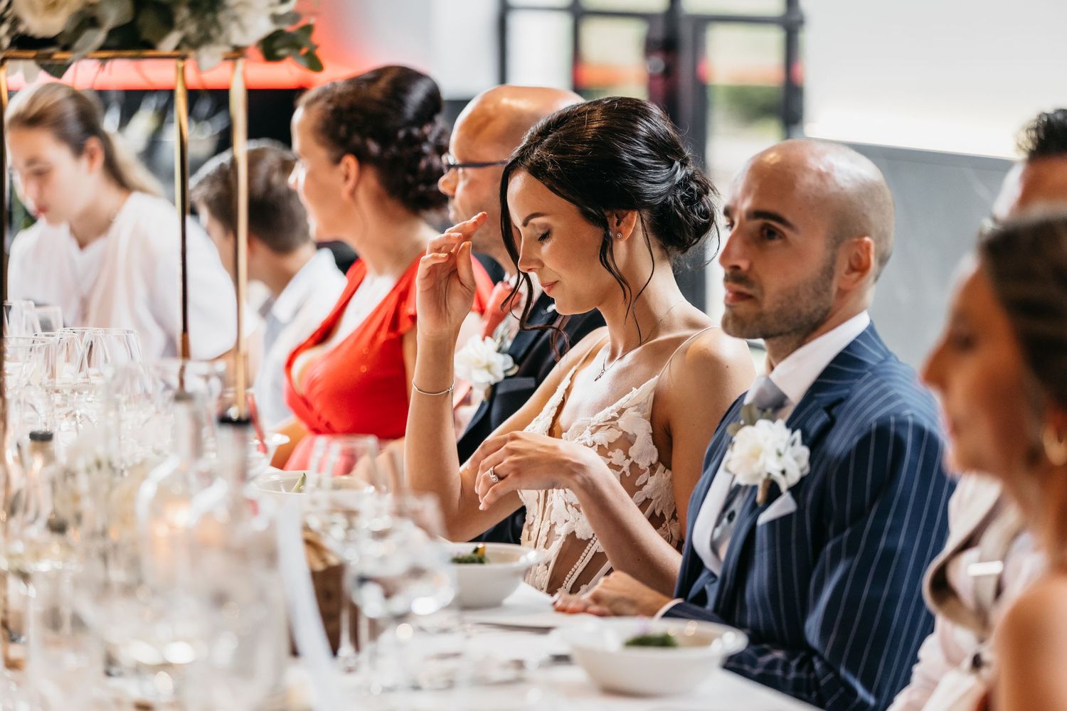 Bride and groom sit attentively at wedding reception dinner table surrounded by guests and glassware.