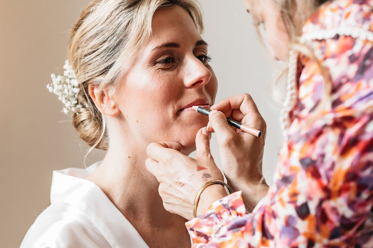 Makeup artist carefully applies lipstick to smiling bride wearing white robe and floral hair piece.