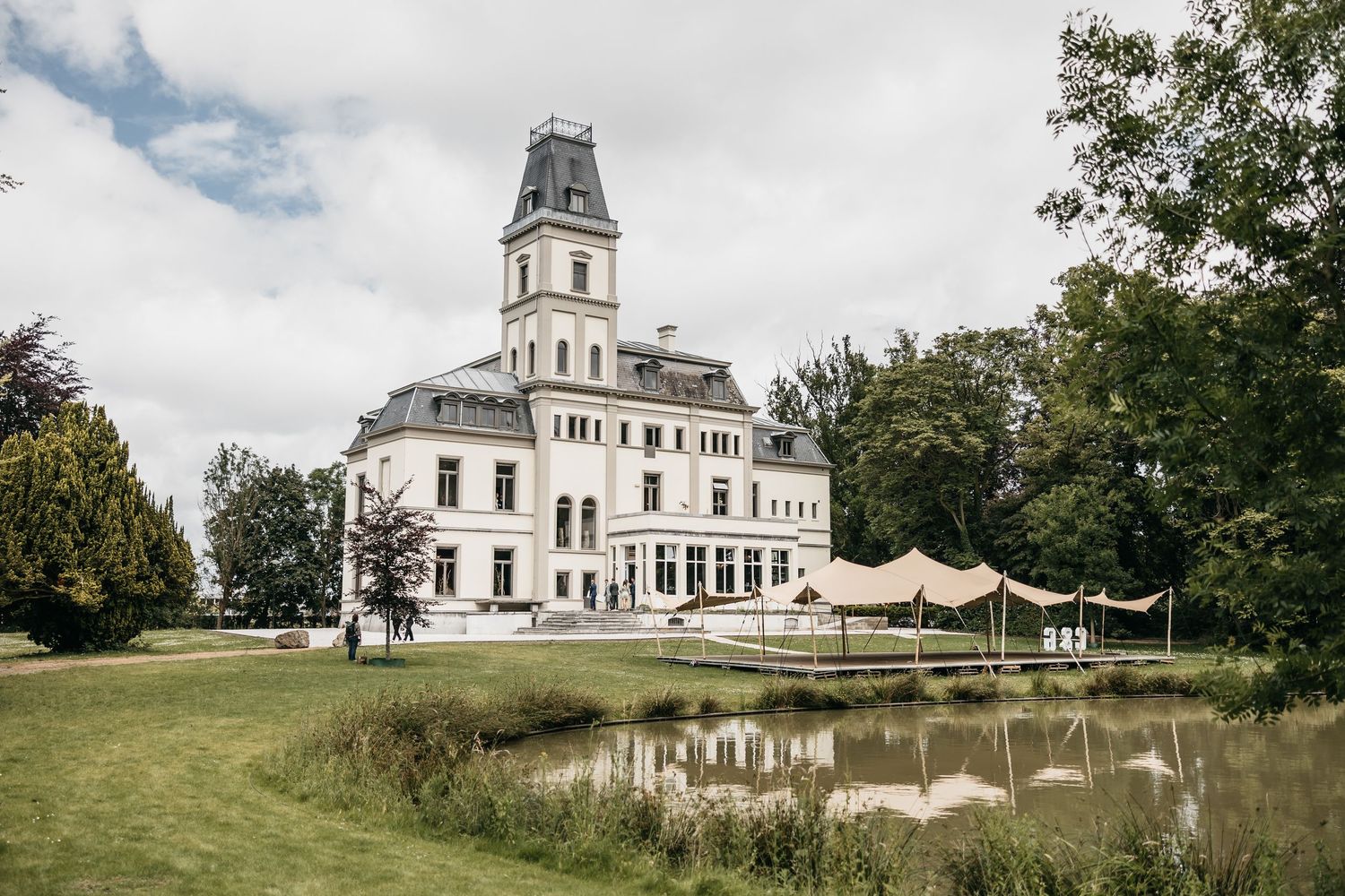 Grand white historic manor house with tower reflected in pond, surrounded by lush gardens and wedding tent.