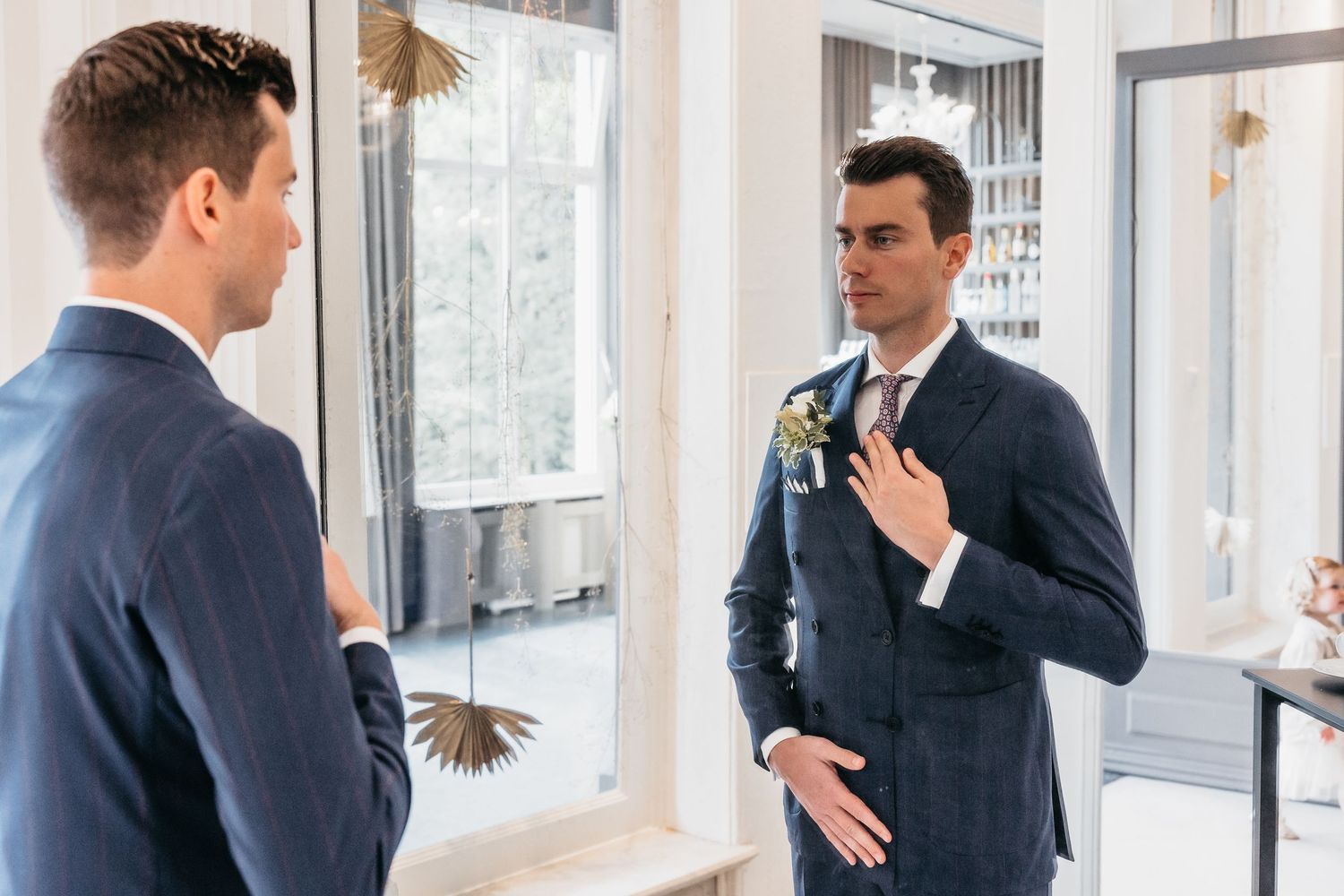 Groom in navy suit checks his boutonniere while looking at his reflection in a mirror before wedding.
