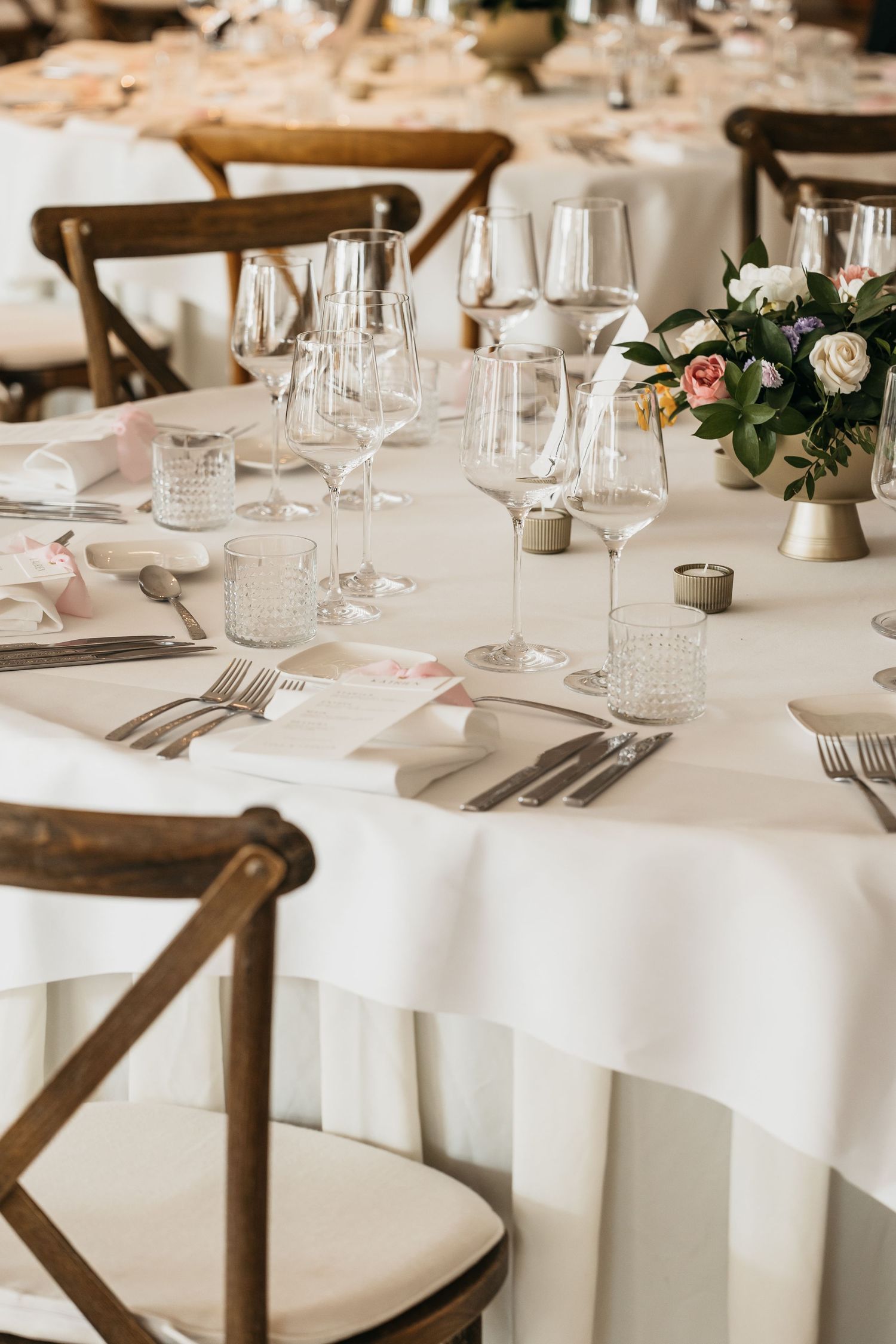 Round wedding reception table with white linen, crystal glasses, floral centerpiece, and wooden chairs.