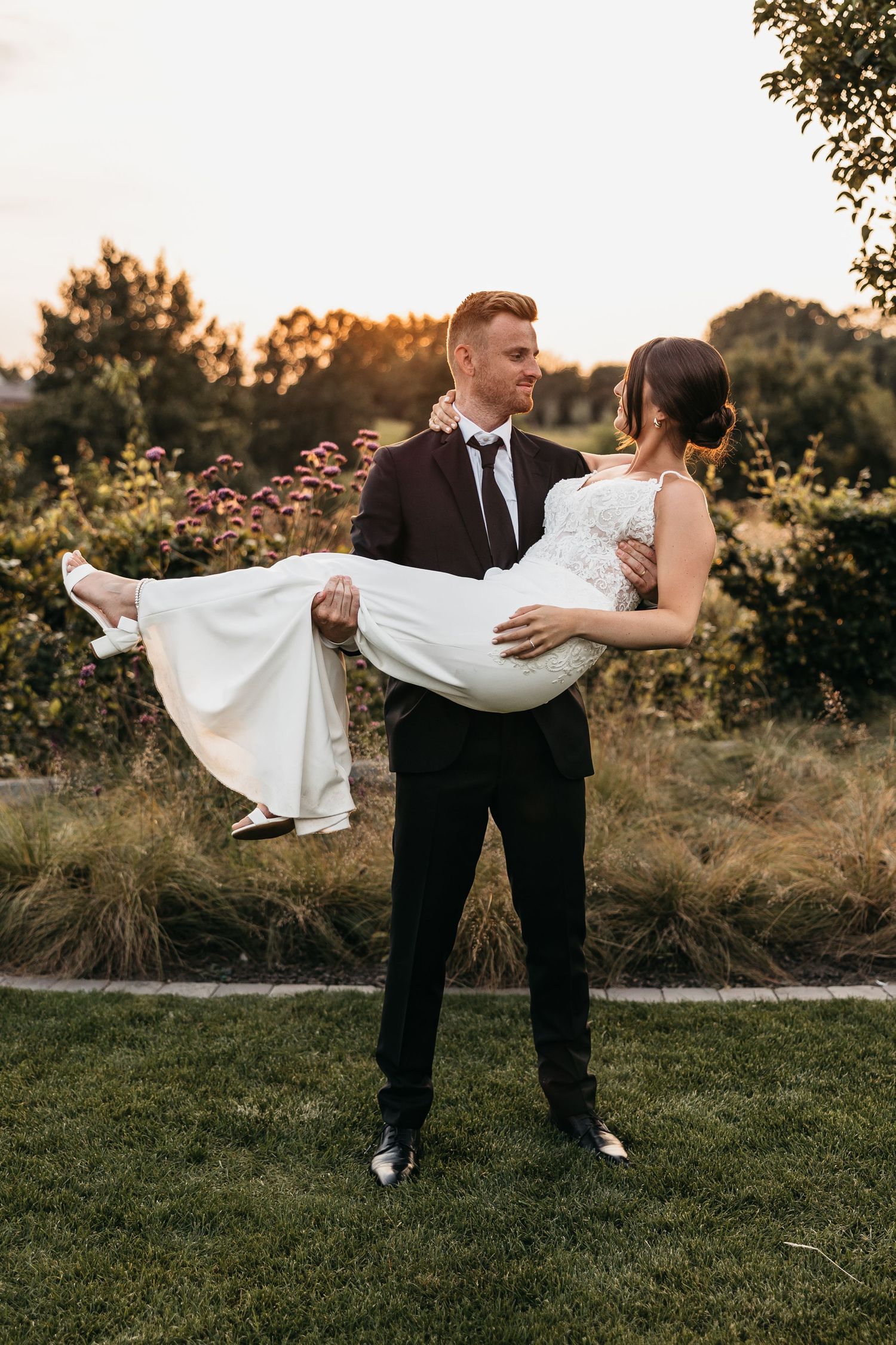 Groom in black tuxedo carries smiling bride in white lace gown in his arms outdoors during a golden sunset.