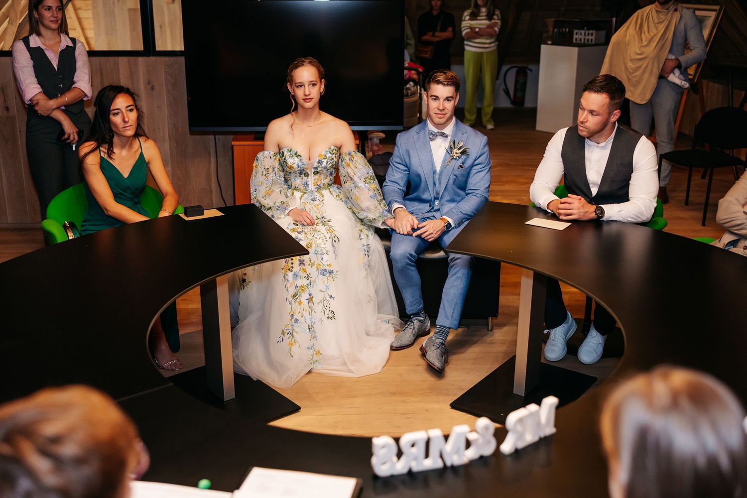 Bride and groom hold hands at circular table during intimate wedding ceremony indoors.