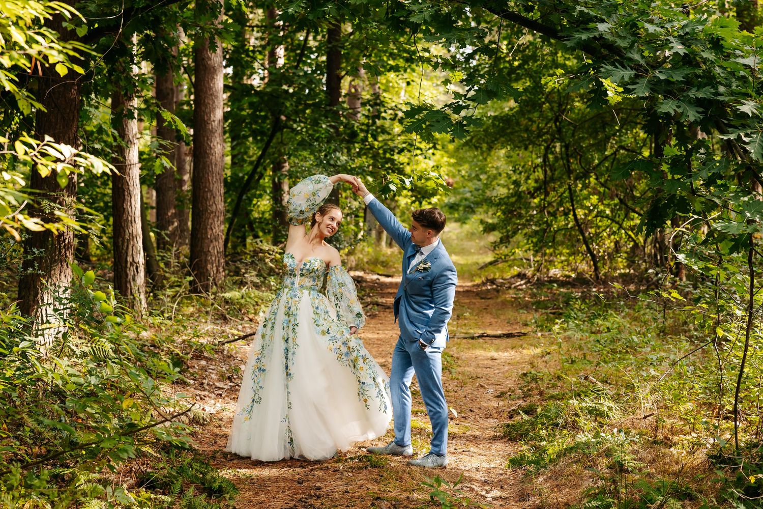 Groom spinning bride in floral gown on scenic woodland path.