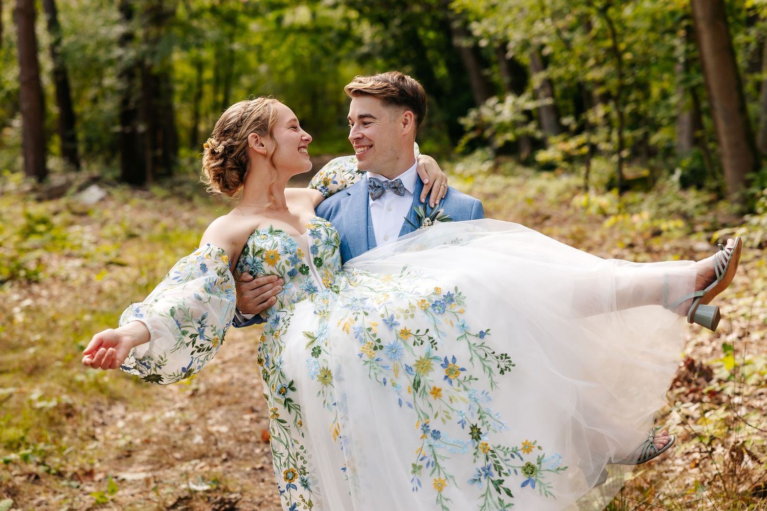 Groom carrying smiling bride in a floral white gown through a sunny woodland forest path.
