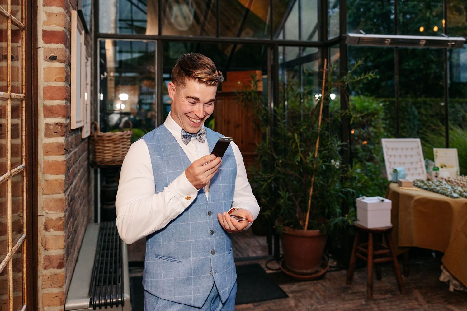 Smiling groom in blue vest and bow tie checking his phone at wedding venue.