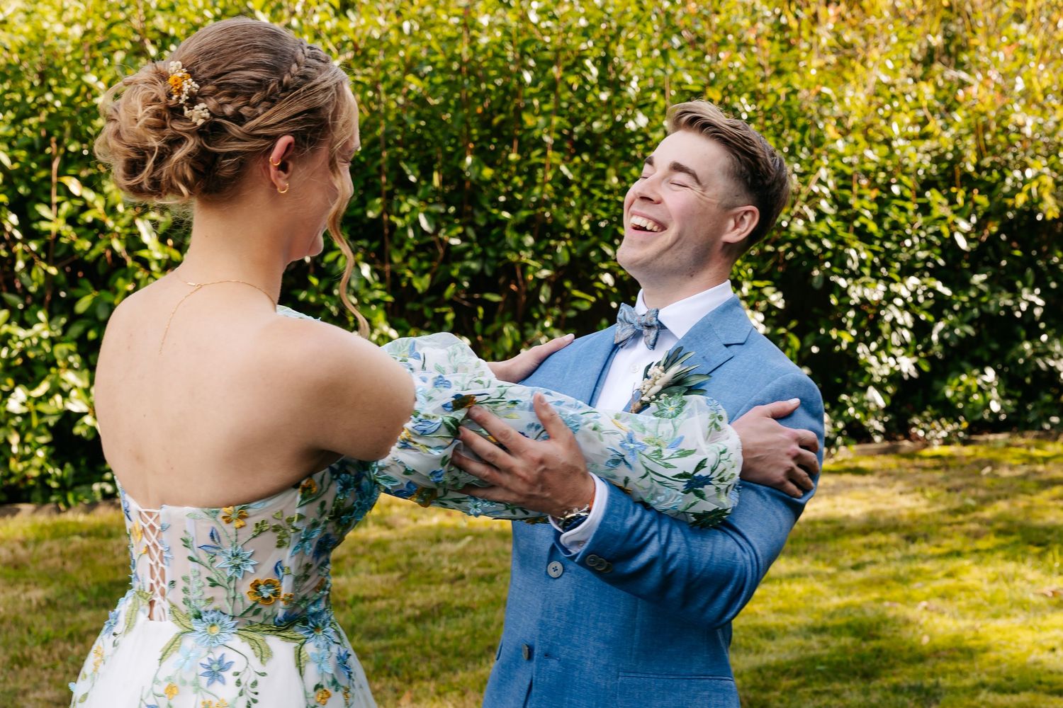 Groom in blue suit laughs with delight while holding bride in floral dress during outdoor first look.