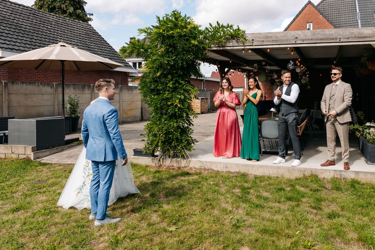 Bride and groom approach cheering wedding guests standing outside sunny garden venue.