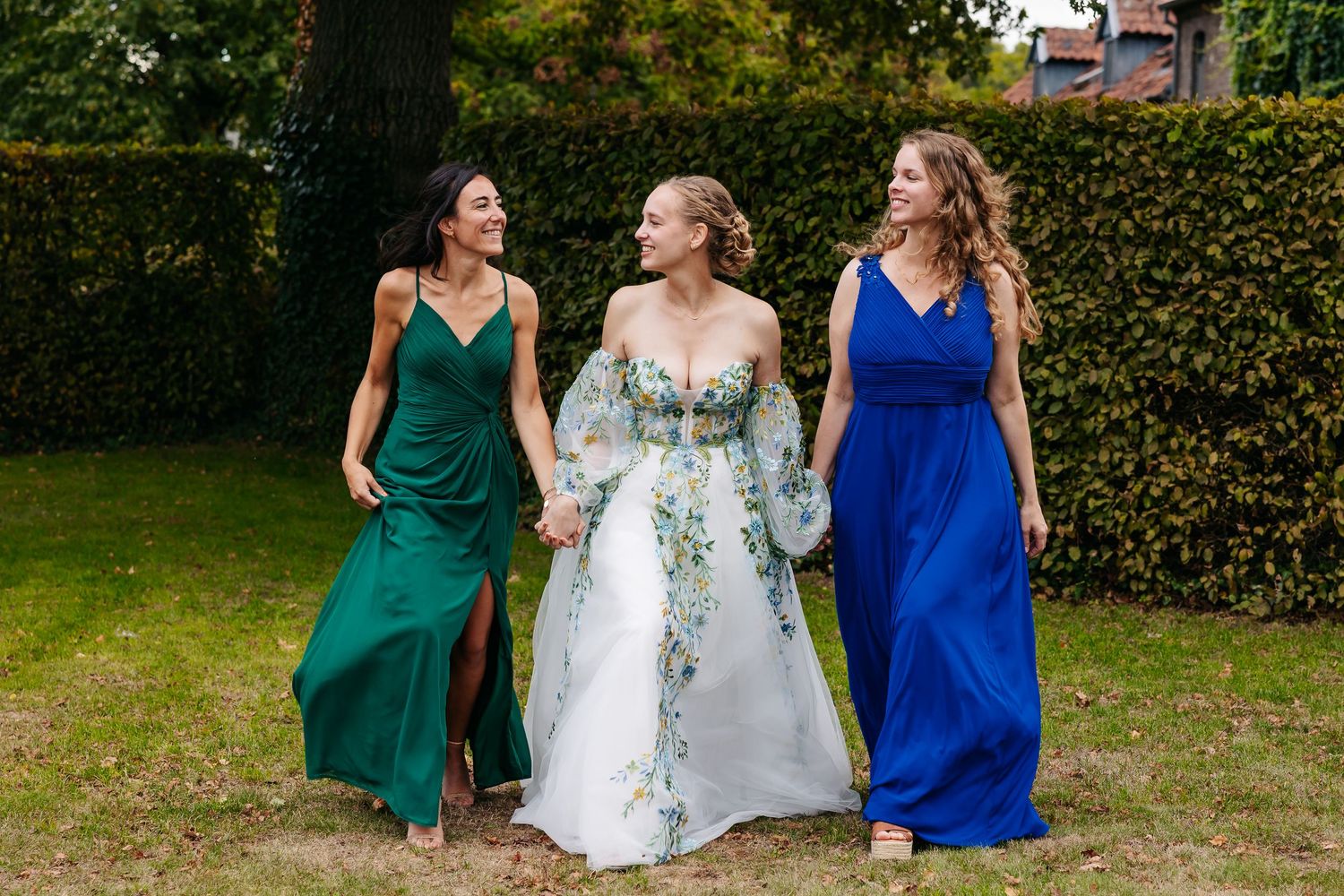 Bride in floral wedding dress walking outdoors with two bridesmaids in green and blue gowns.