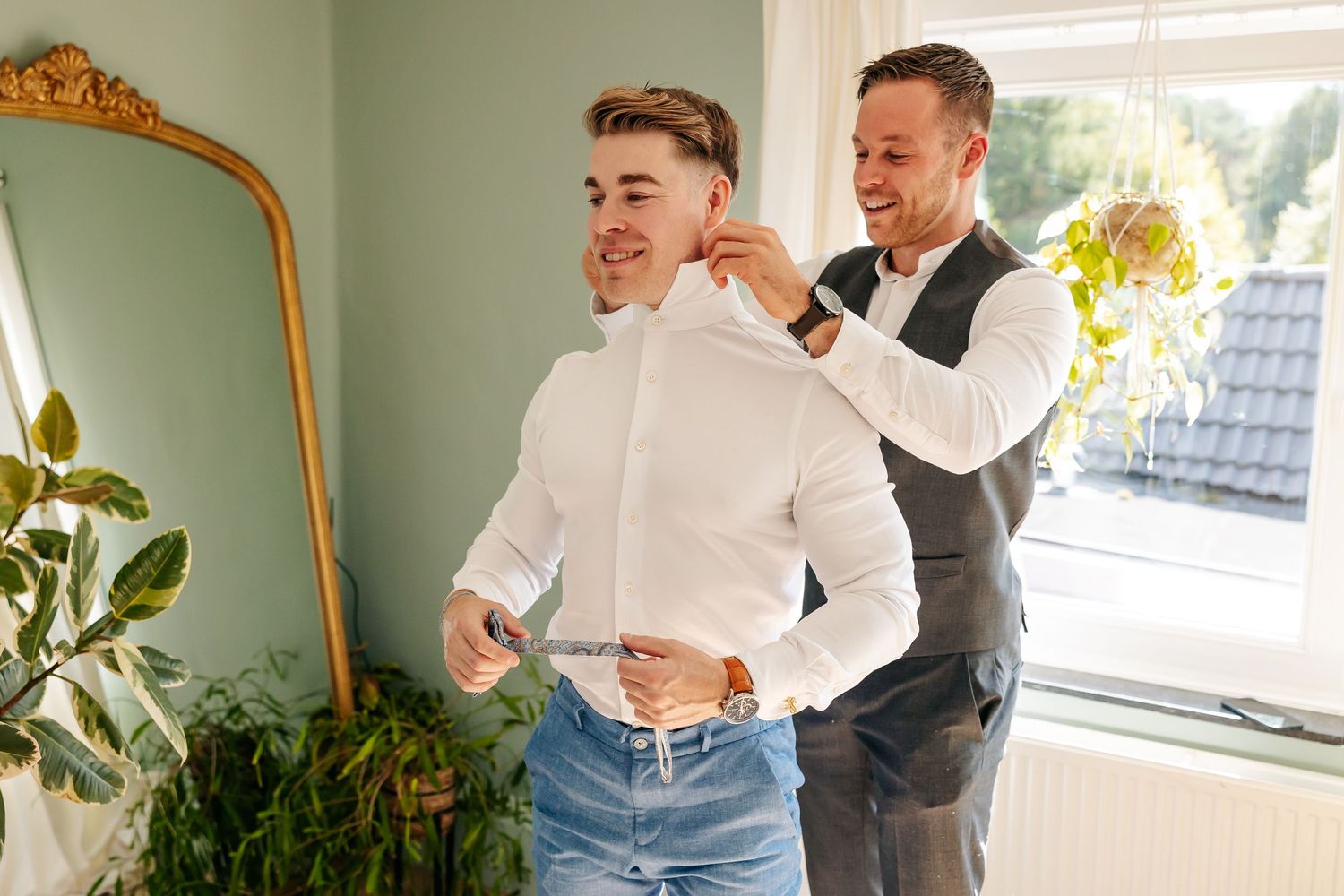 Groomsman helping groom adjust collar while getting ready for wedding in bright room.