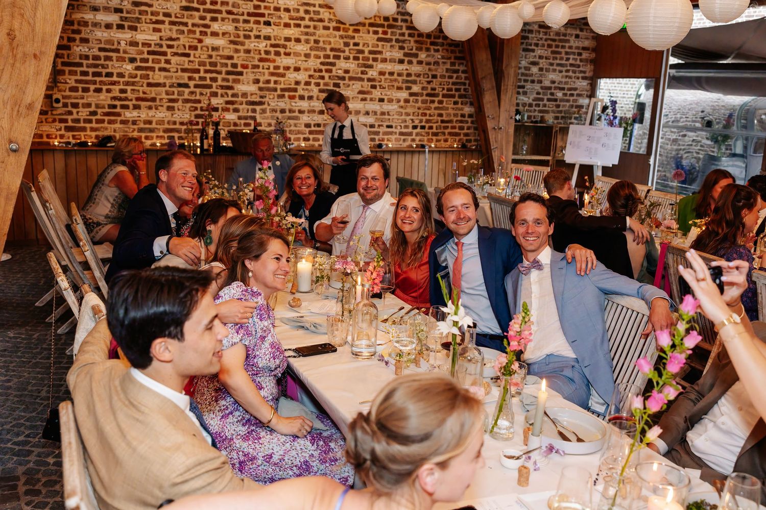 Wedding guests smiling and celebrating at a decorated reception table in a rustic brick venue.