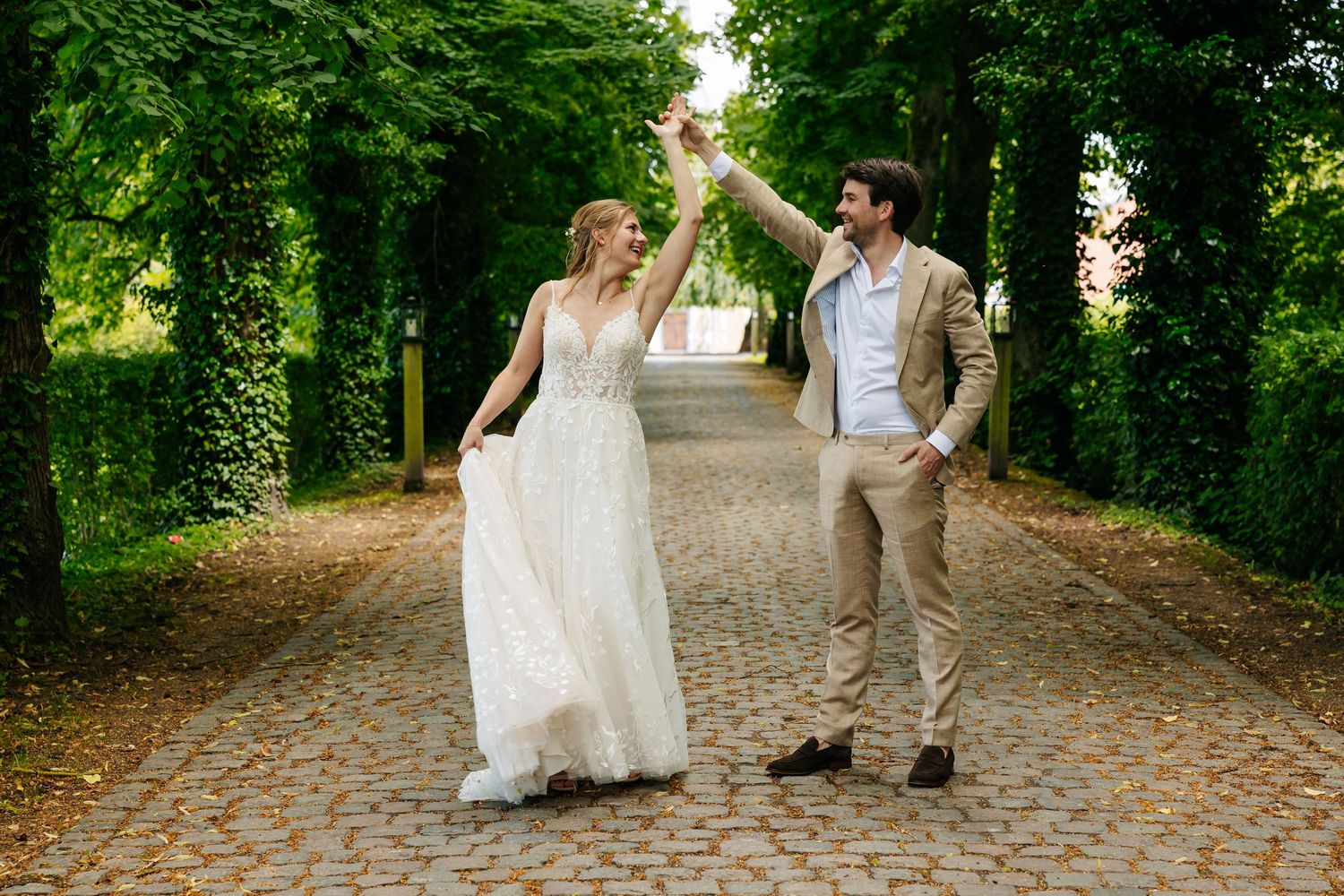 Bride and groom dancing joyfully on tree-lined cobblestone path surrounded by lush green foliage.