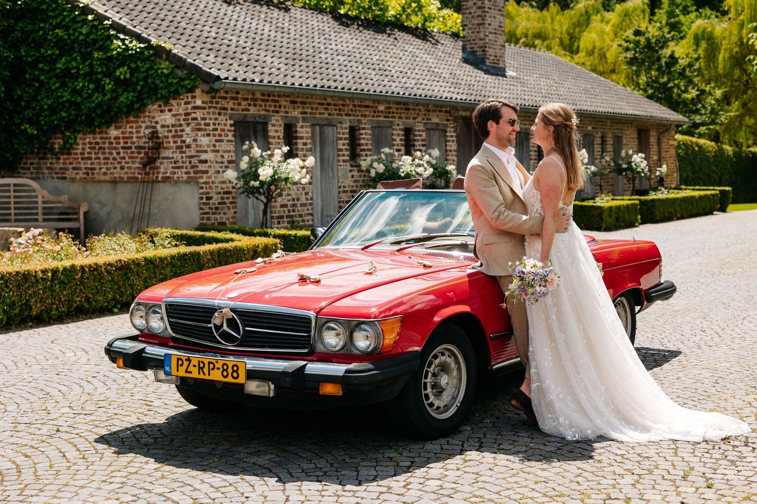 Bride and groom posing beside classic red Mercedes convertible at rustic wedding venue.