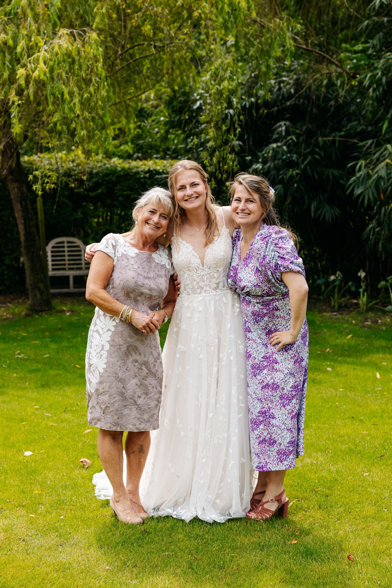 Bride in white lace gown posing with two women in garden on wedding day.