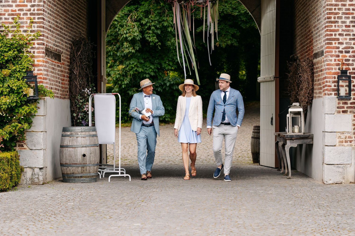 Three stylish wedding guests walking through a rustic brick archway decorated with colorful ribbons.