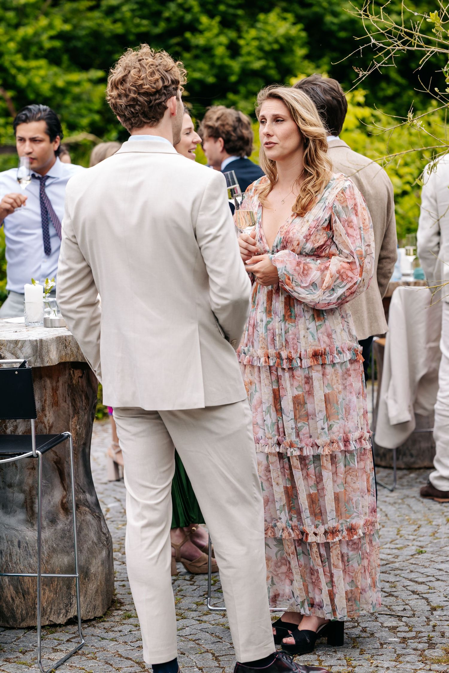 Wedding guest in floral dress converses with man in beige suit during outdoor cocktail reception.