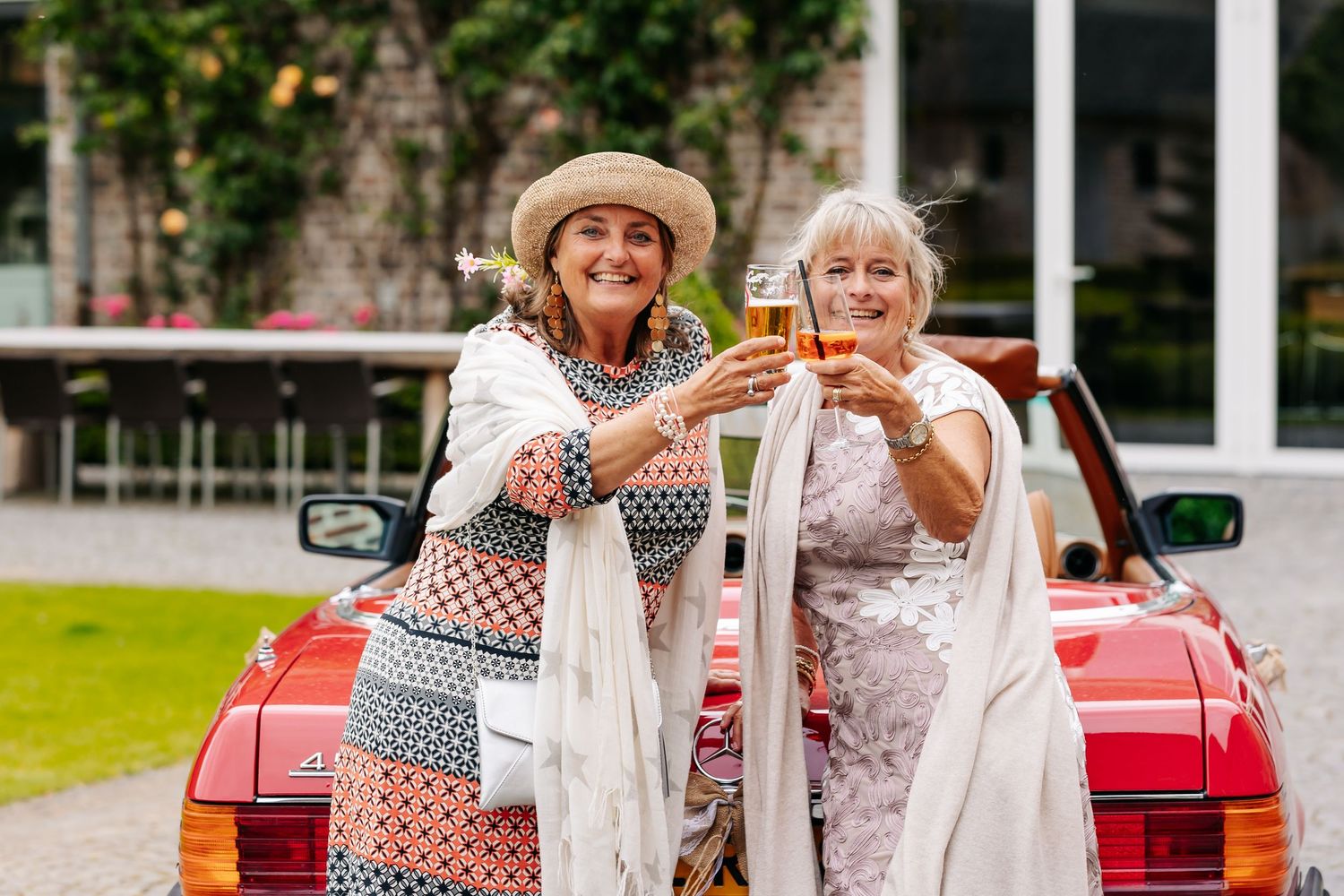 Two smiling women toast with champagne glasses leaning against a red vintage convertible car outdoors.