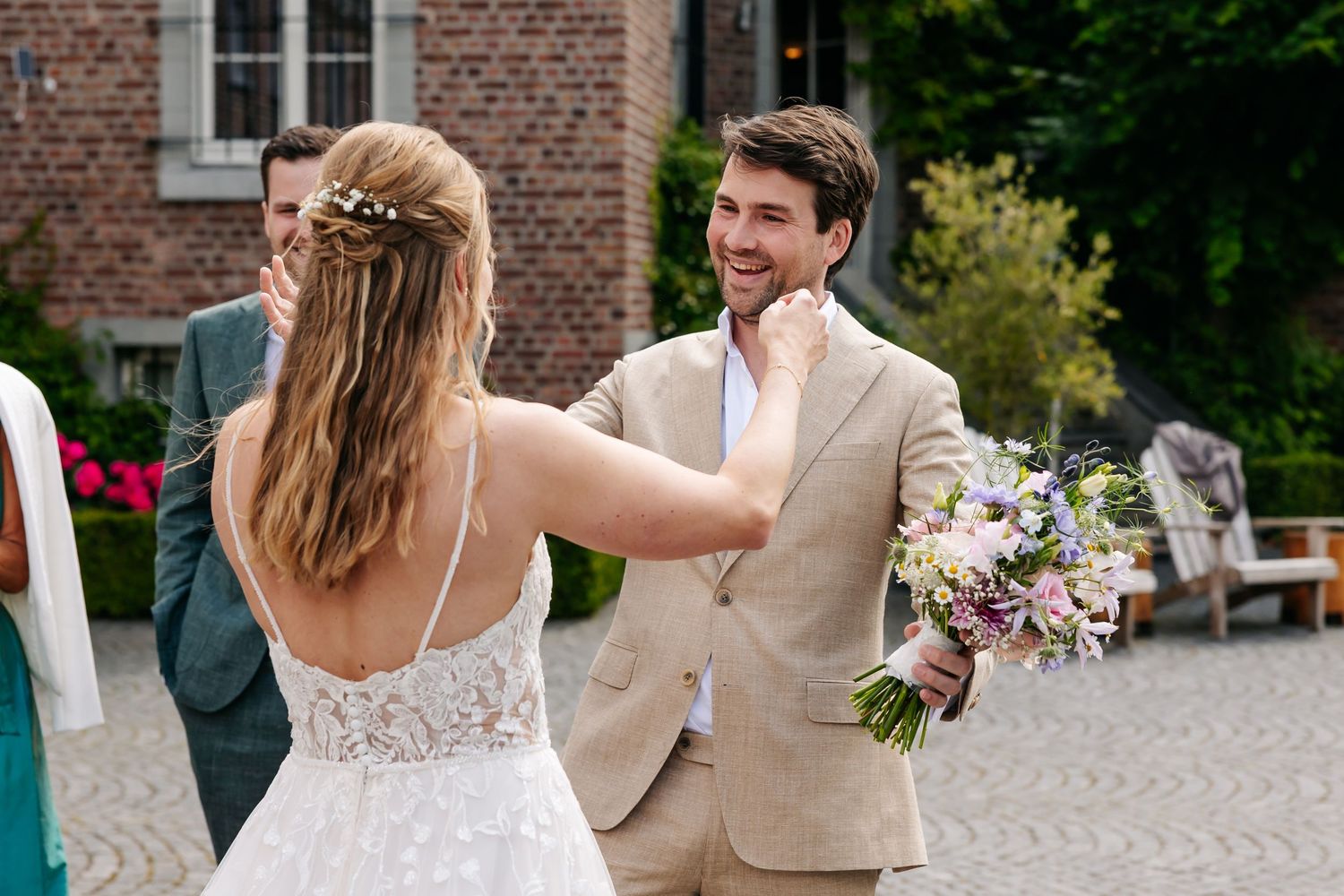 Groom in beige suit holding bouquet smiling at bride during outdoor wedding ceremony.