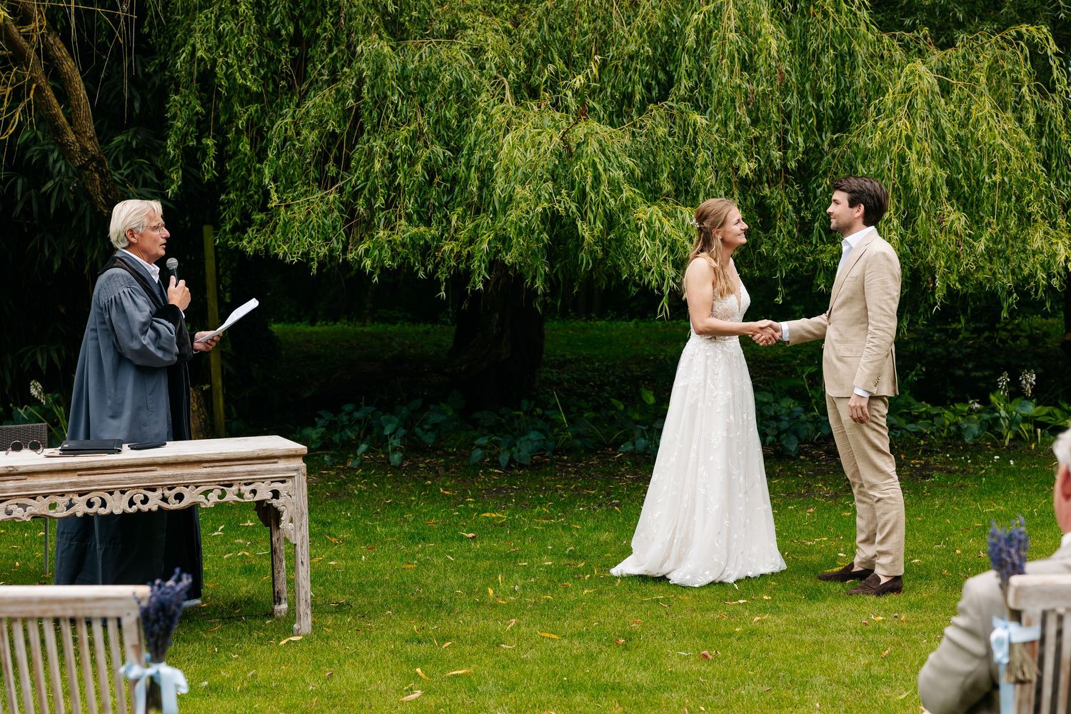 Officiant conducting outdoor wedding ceremony under willow tree as couple holds hands on lawn.