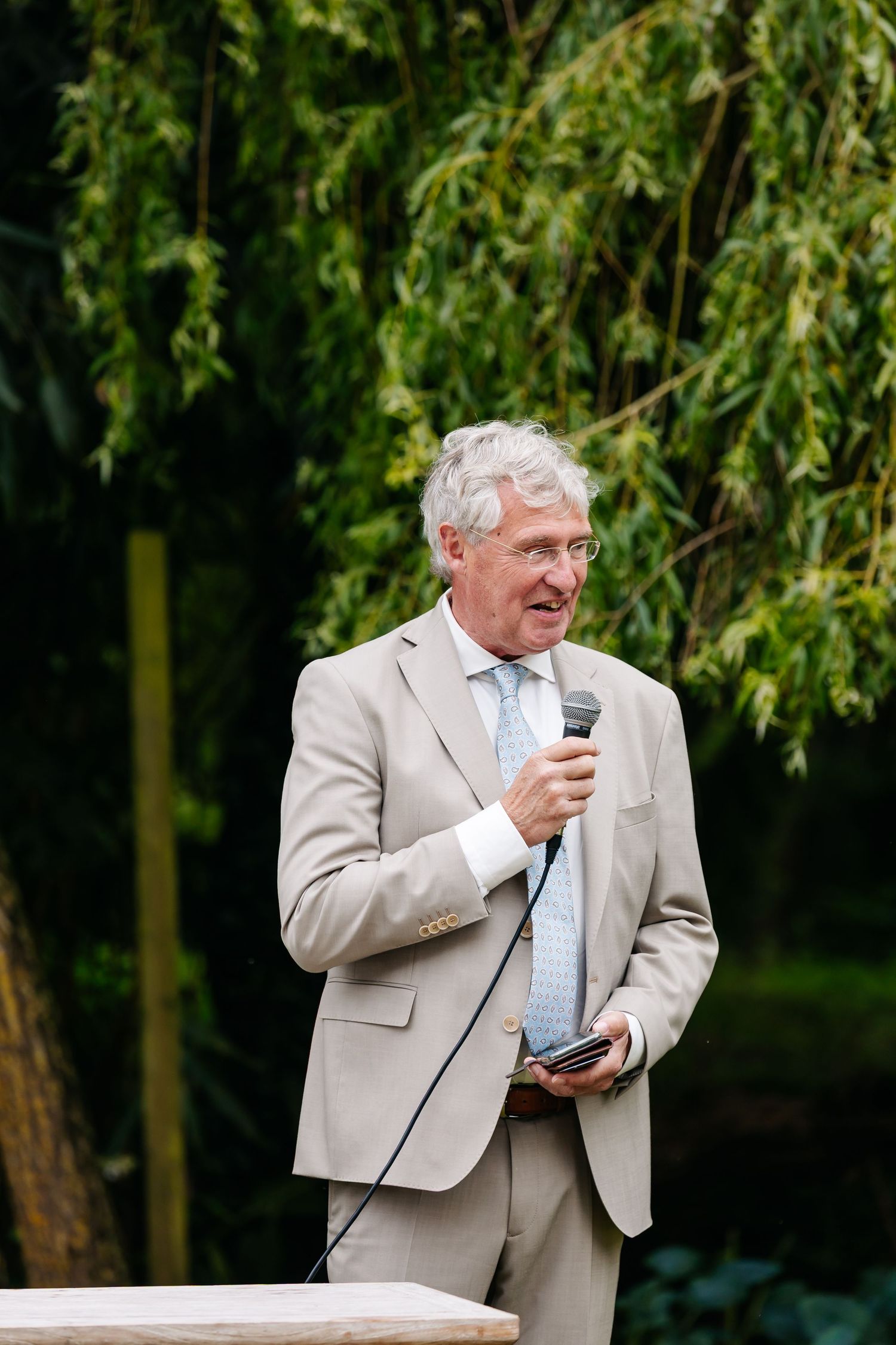 Elegant older man in beige suit speaks into microphone at outdoor garden event.