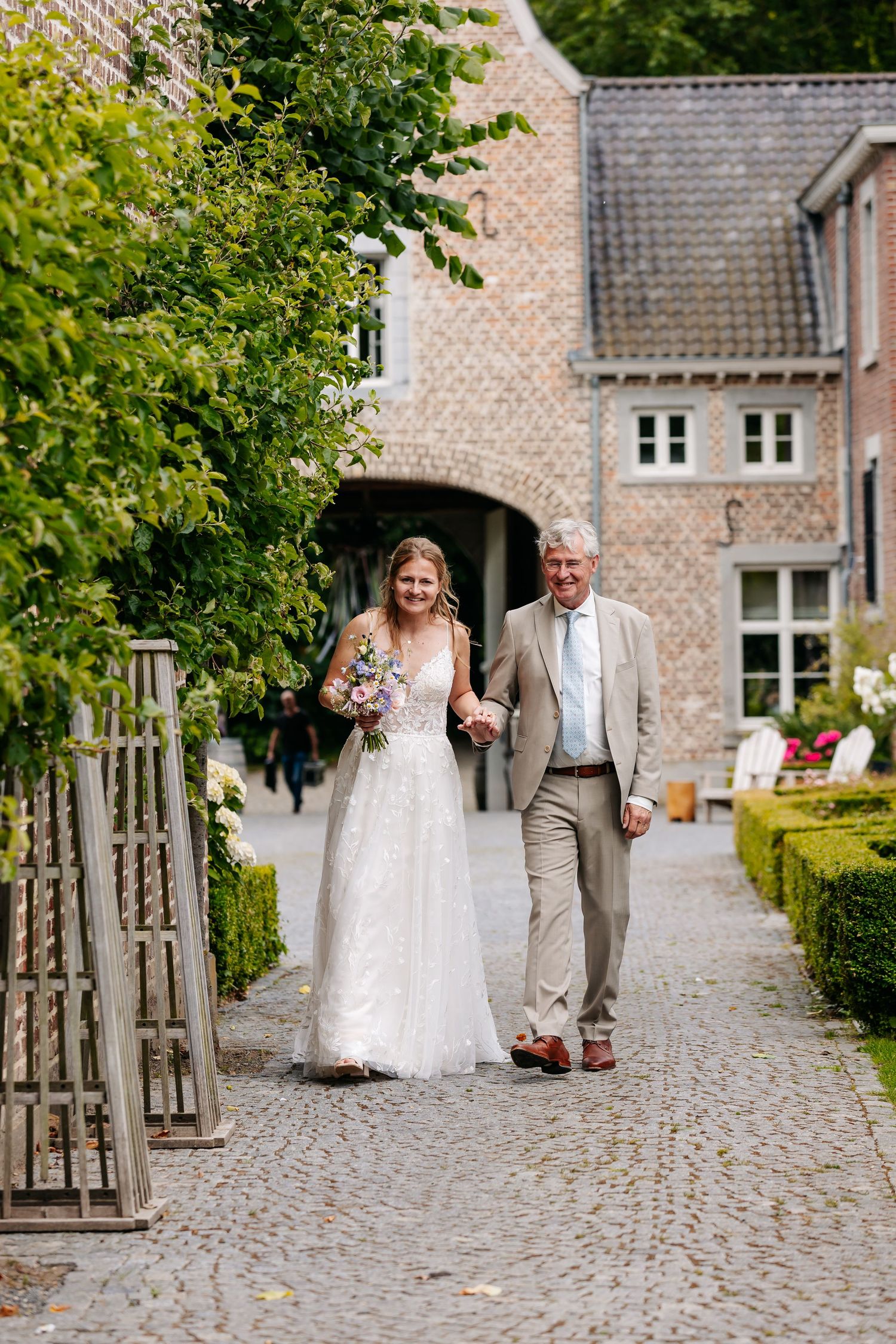 Smiling bride in lace gown holding a bouquet walks with an older man in beige suit along a cobblestone path.