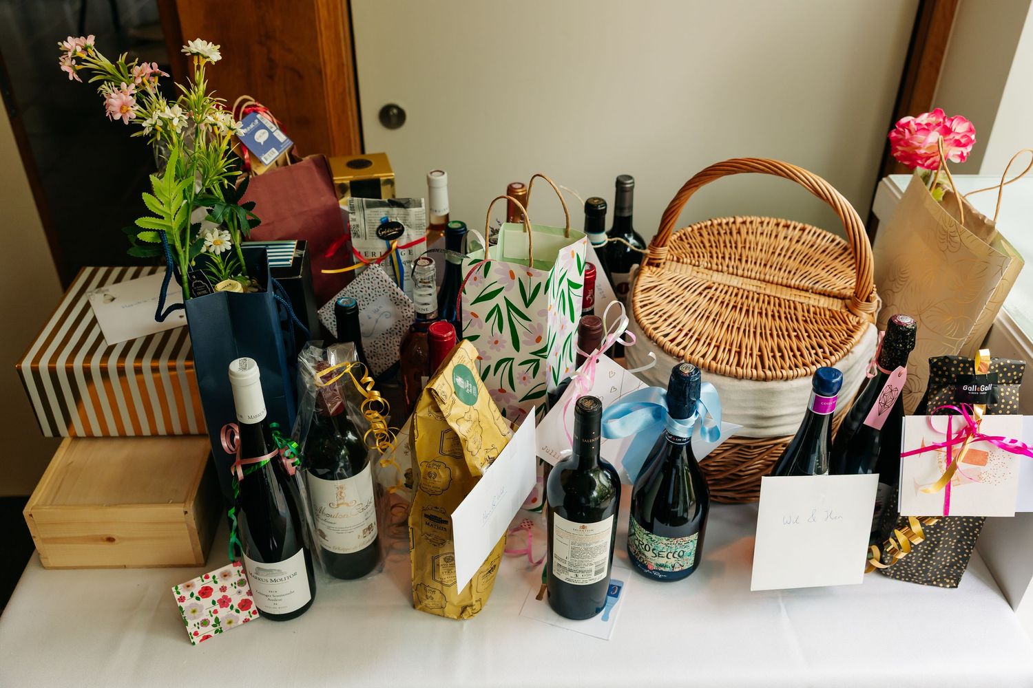 Wedding gift table displaying wine bottles, wicker basket, flowers, and wrapped presents at reception.