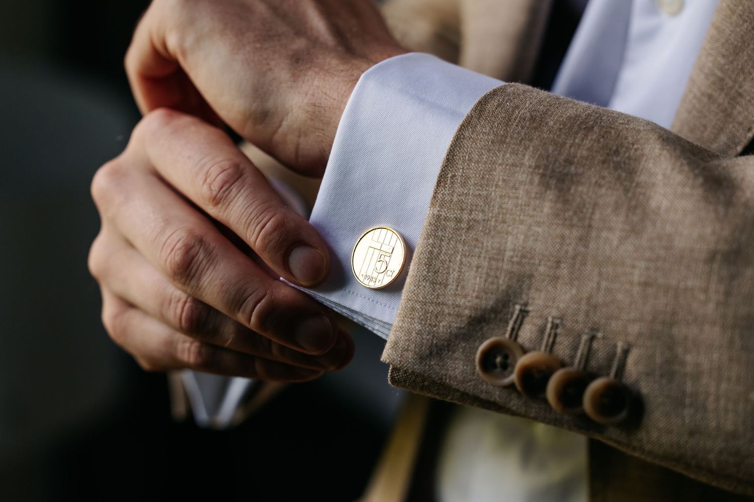 Close-up of groom fastening gold cufflink on white dress shirt with tan suit jacket.
