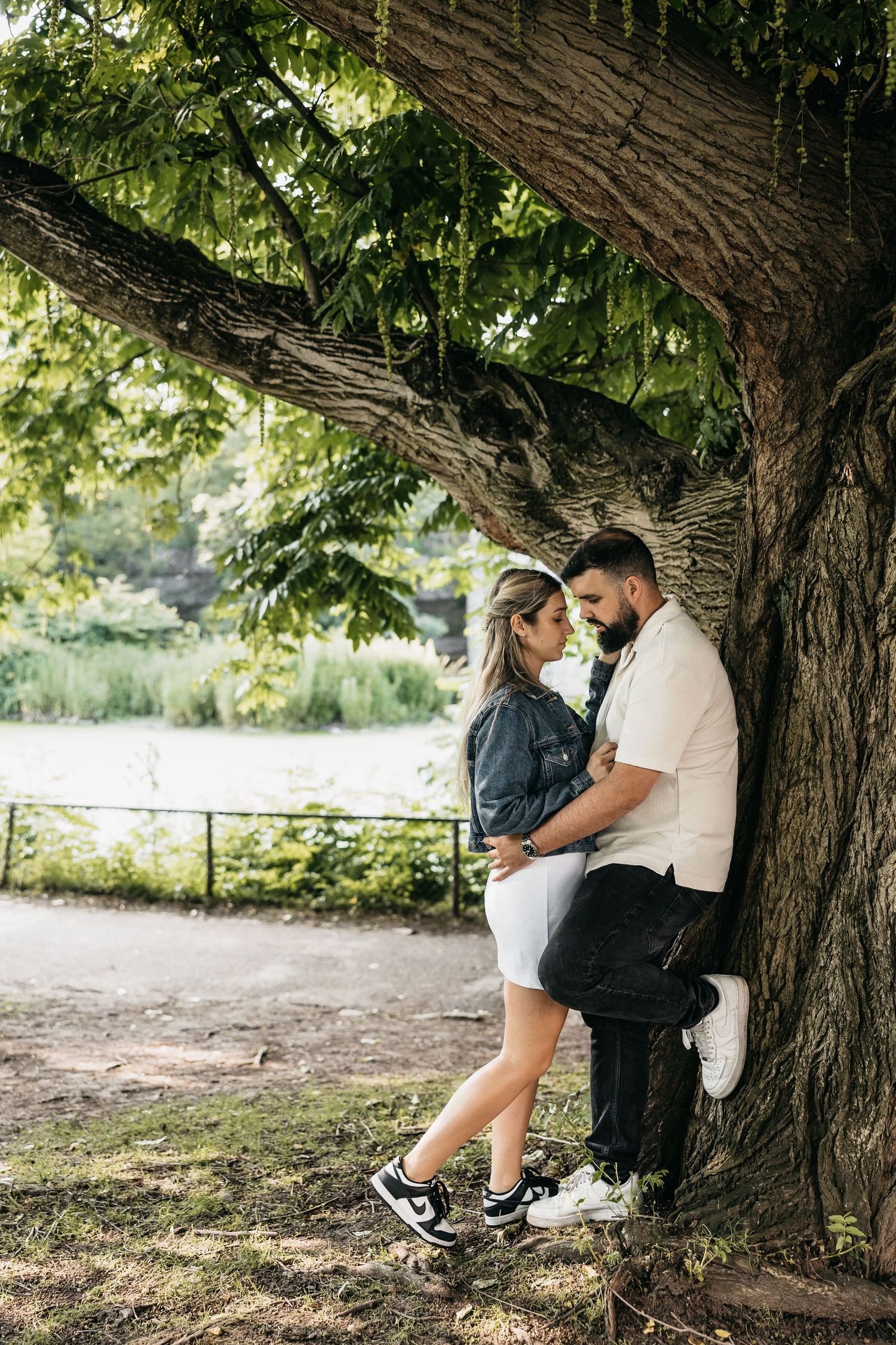 Couple embracing romantically under a large tree in a lush green park setting.