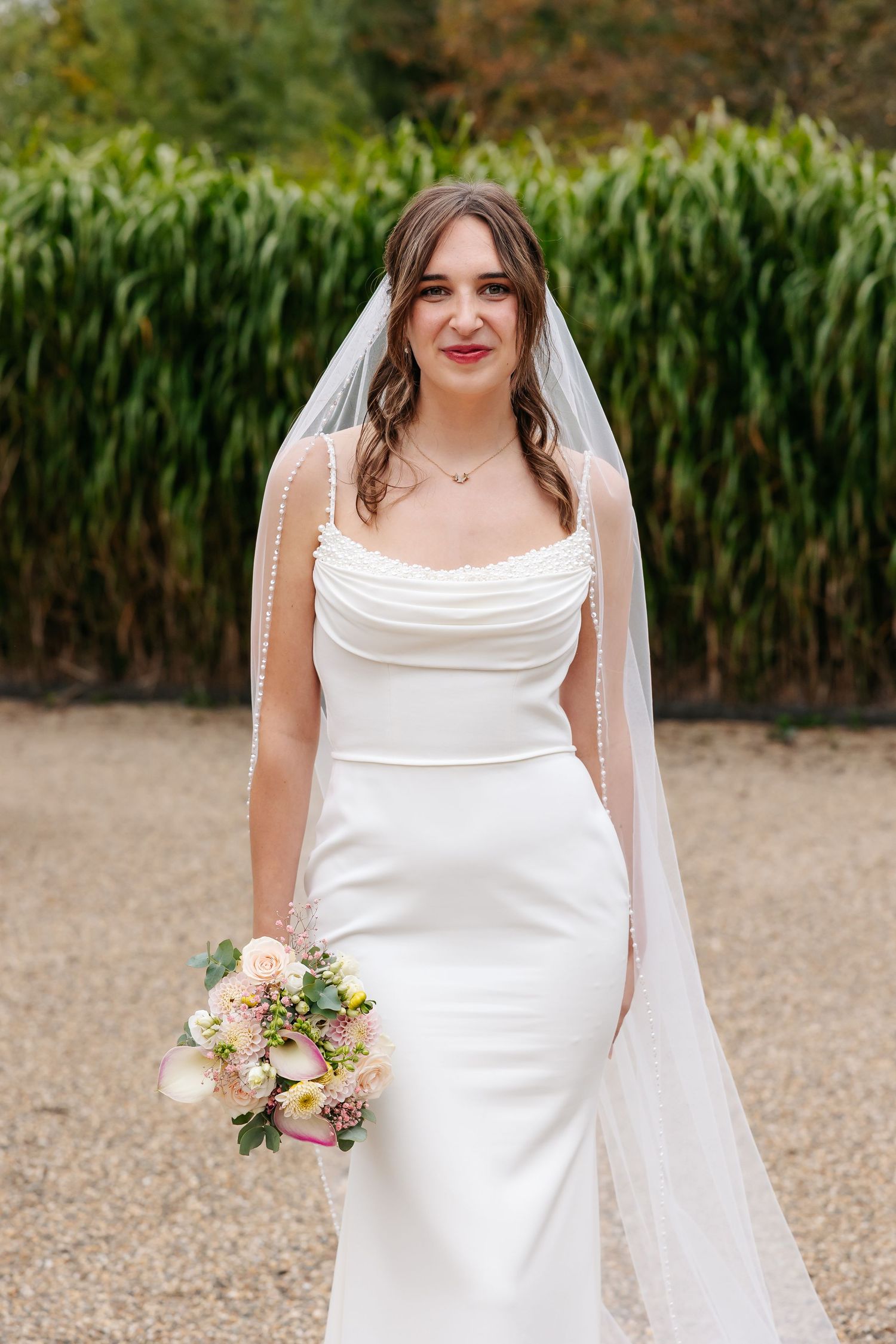 Elegant bride in white gown and veil holds pastel bouquet outdoors.