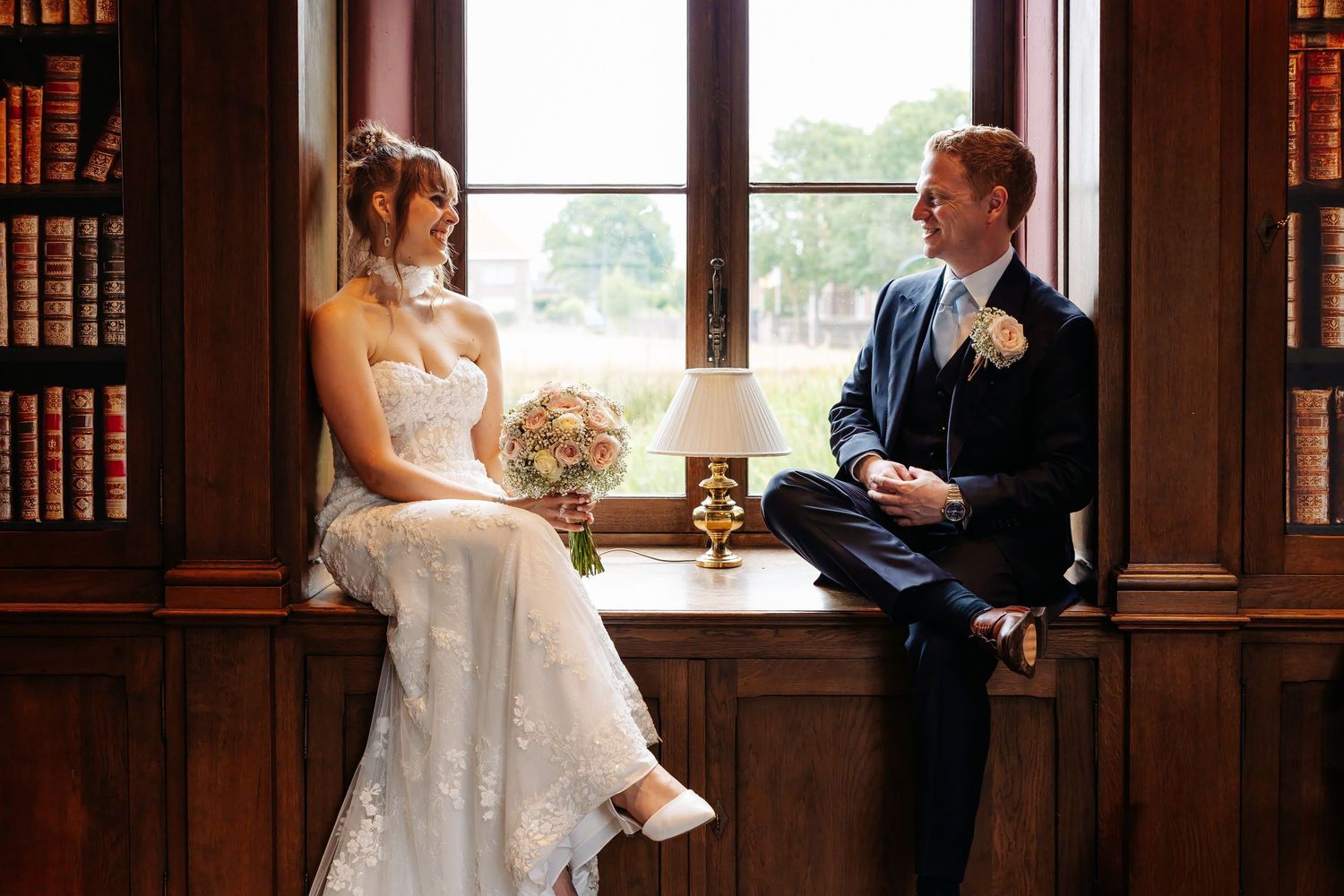 Bride and groom sit together on a window seat in an elegant wood-paneled library filled with vintage books.