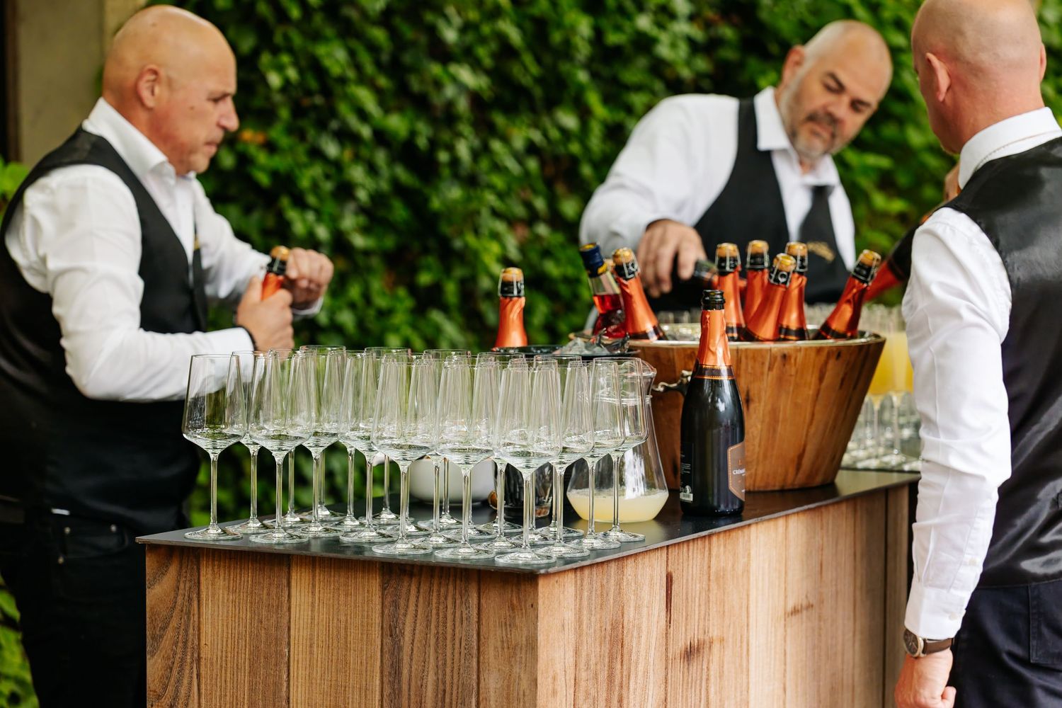 Three bartenders prepare champagne flutes and bottles at a wooden bar station surrounded by lush green foliage.