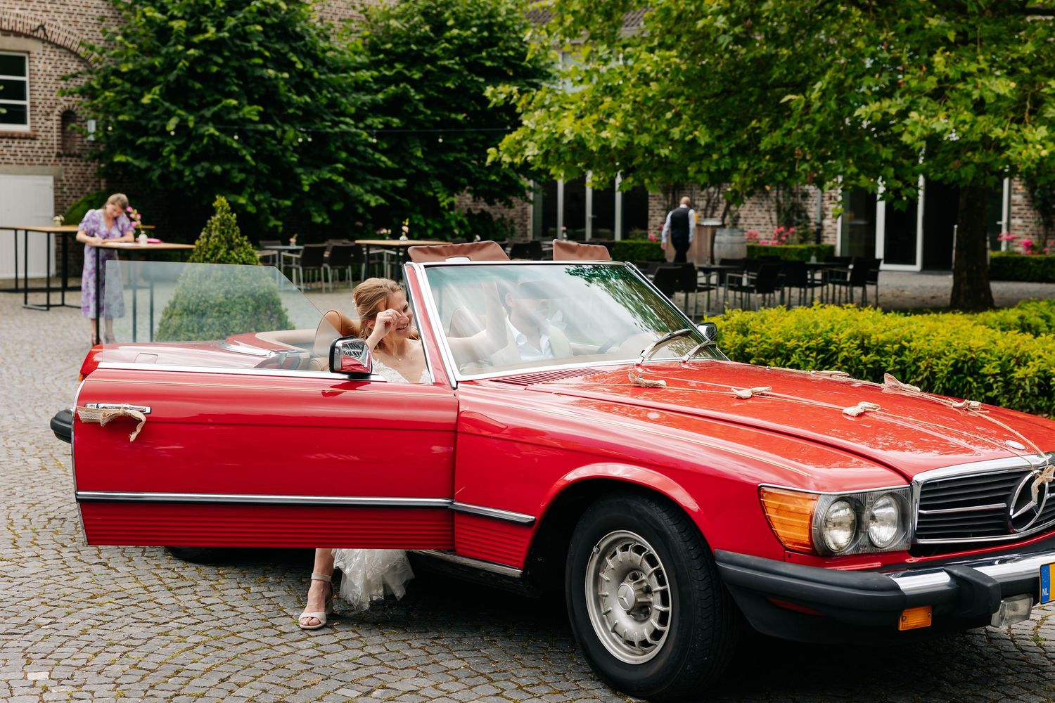 Bride in white gown stepping into a classic red Mercedes-Benz convertible at elegant wedding venue.
