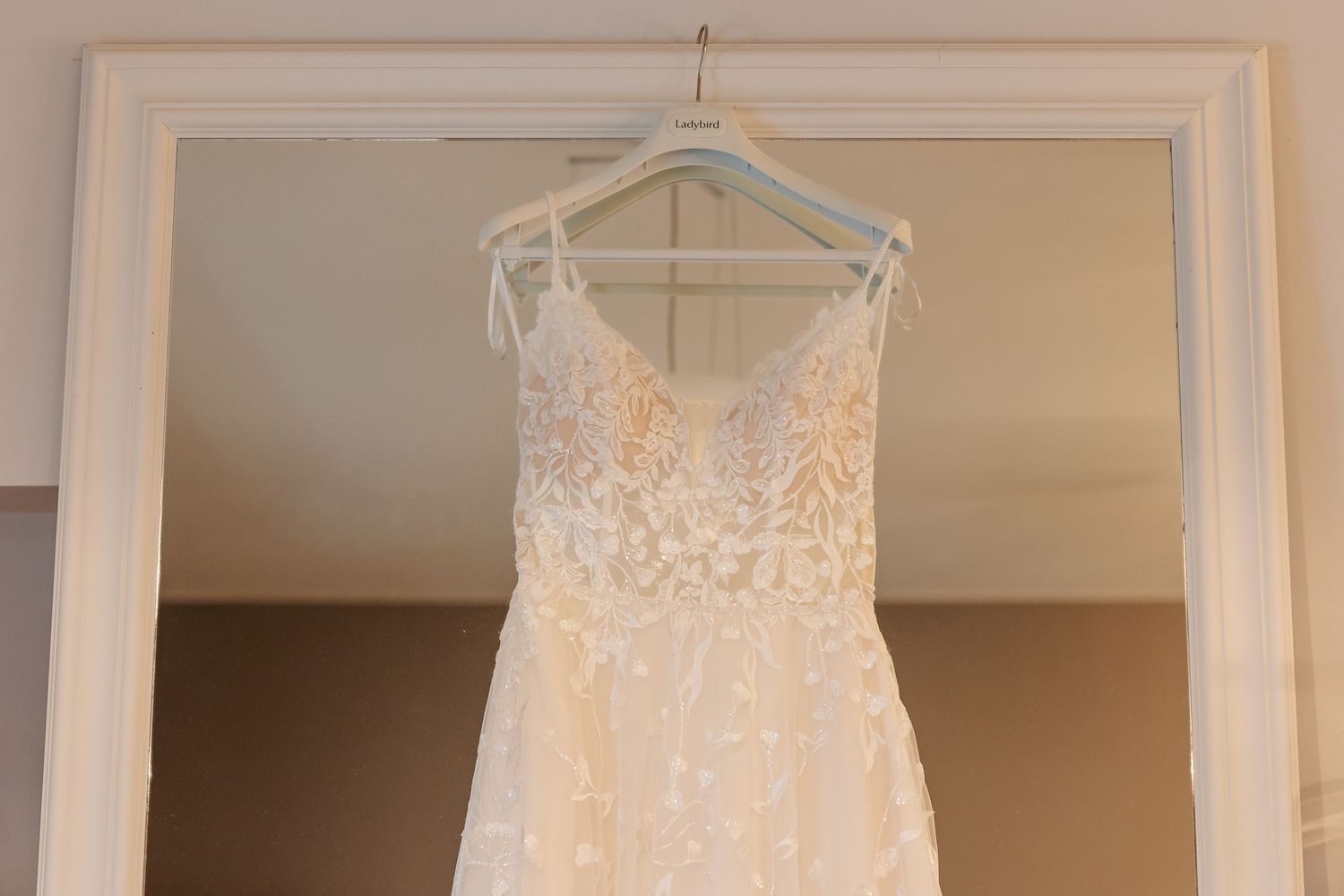 Elegant lace wedding dress with V-neckline hanging on a mirror frame, featuring floral embroidery and spaghetti straps.