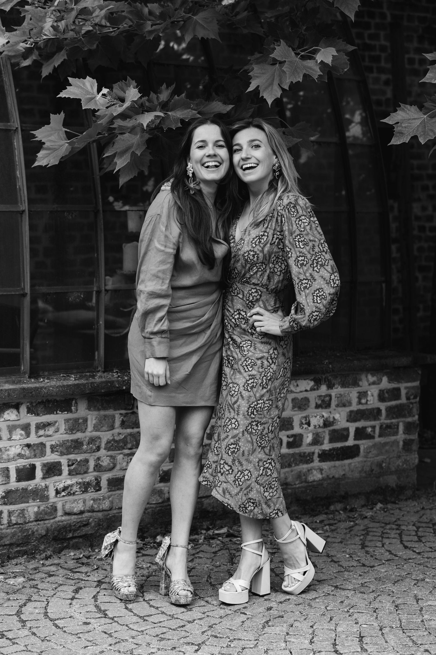 Two elegantly dressed women laughing together in black and white photo against a brick wall backdrop.