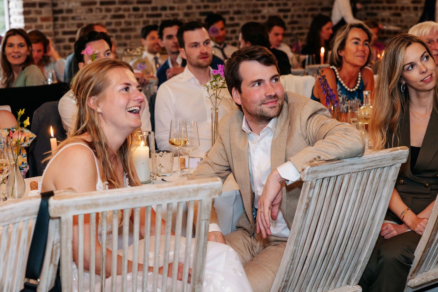 Smiling wedding guests enjoy reception dinner in elegant rustic barn with candlelit tables.
