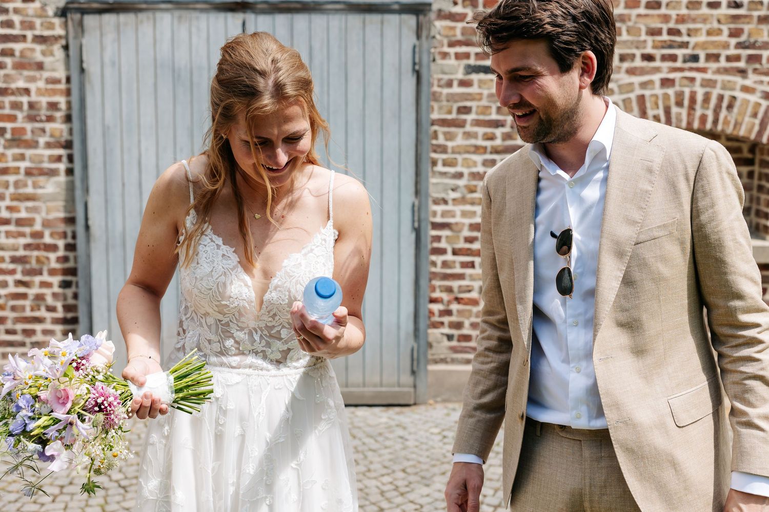 Bride in lace wedding dress holding bouquet and blue item walking alongside groom in beige suit near brick wall.