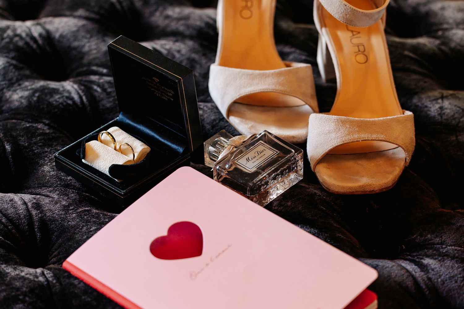 Wedding flat lay with gold rings, perfume bottle, pink heart notebook, and nude heeled sandals.