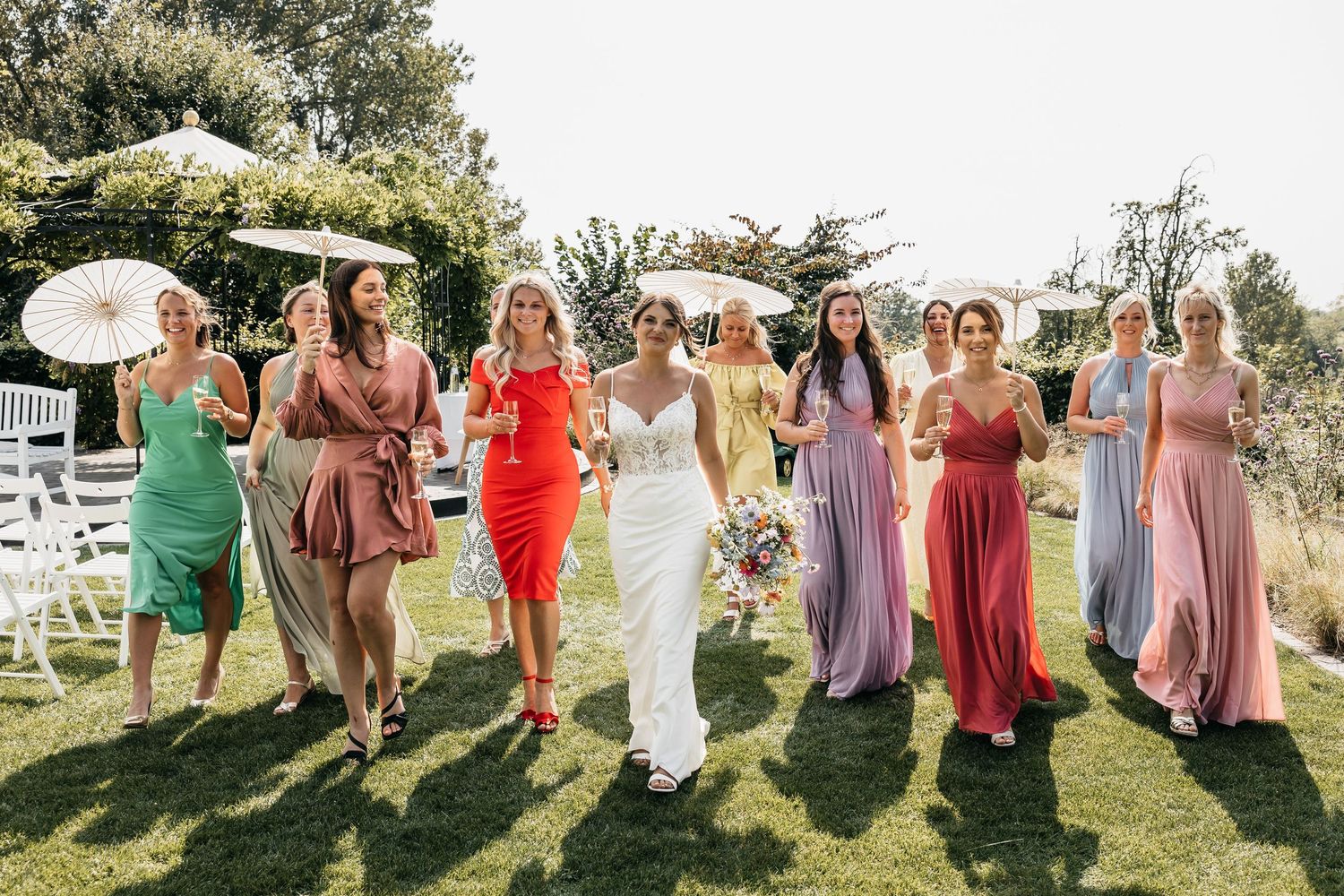 Bride in white gown strolling with bridesmaids in vibrant multicolored dresses across sunny garden.