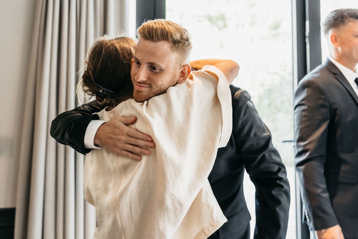 Groom in black suit sharing an emotional hug with a guest during wedding preparations indoors.