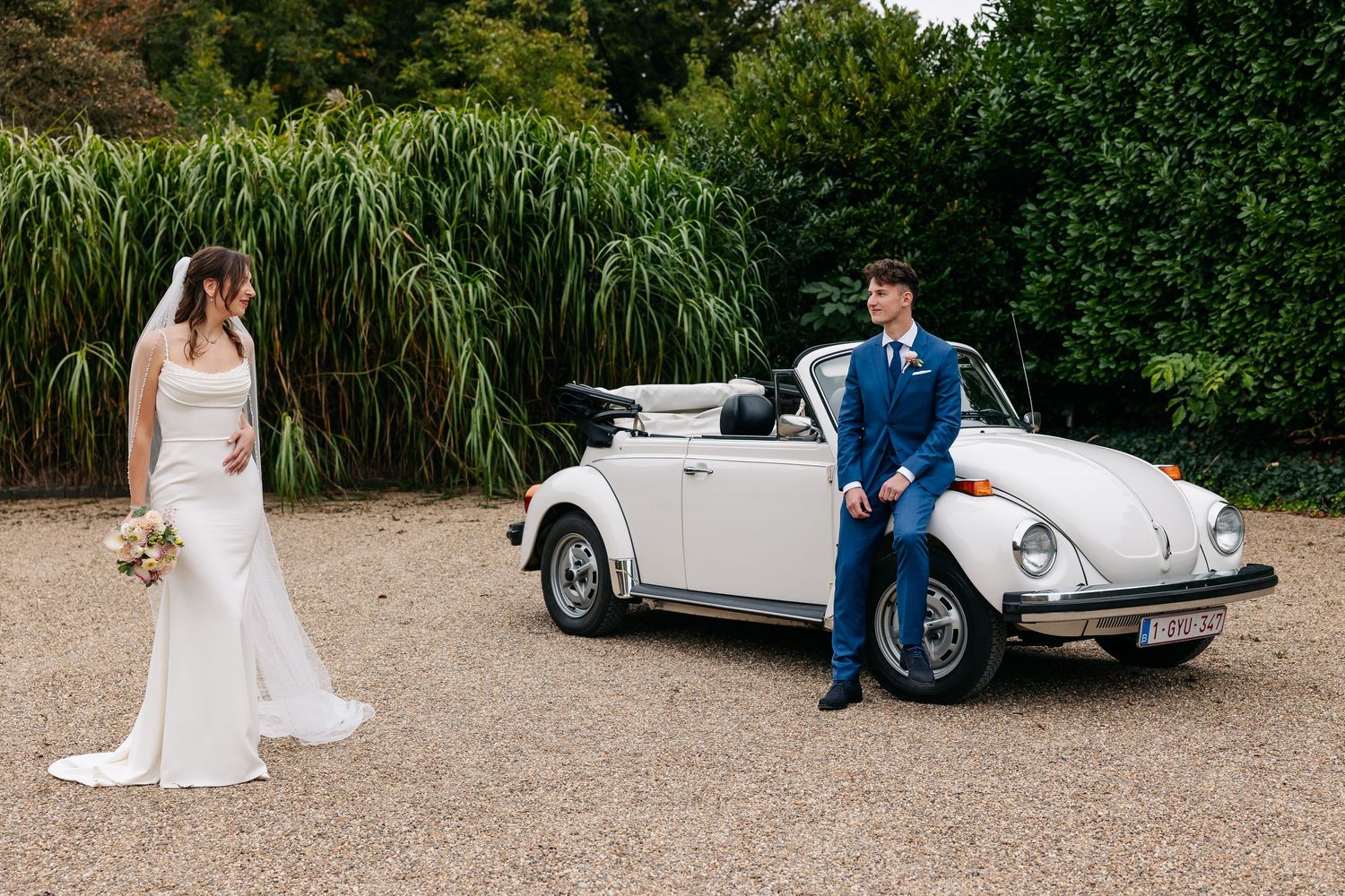 Bride and groom pose beside a classic white VW Beetle convertible on their wedding day.