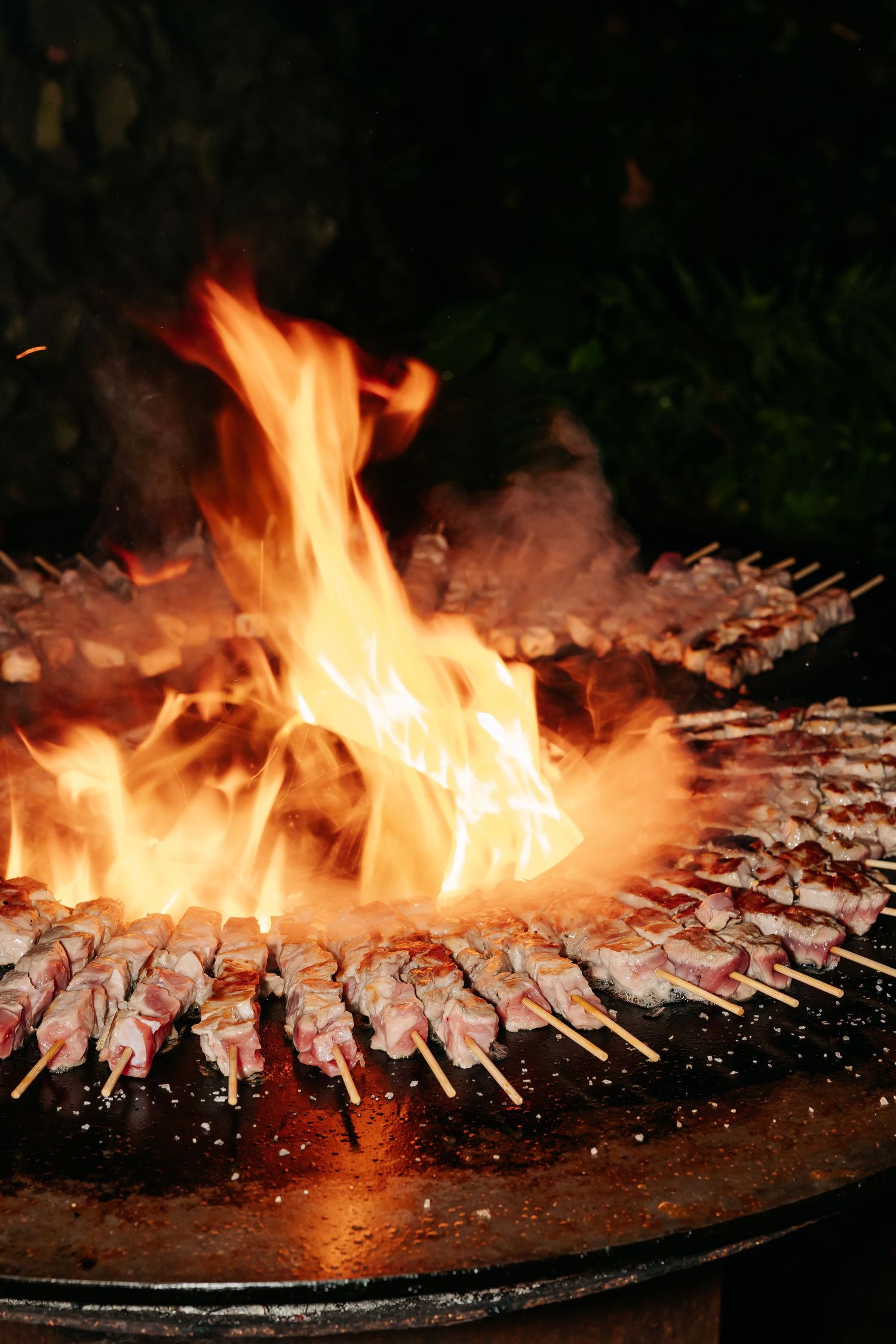 Meat skewers grilling over open flames on a large round BBQ.