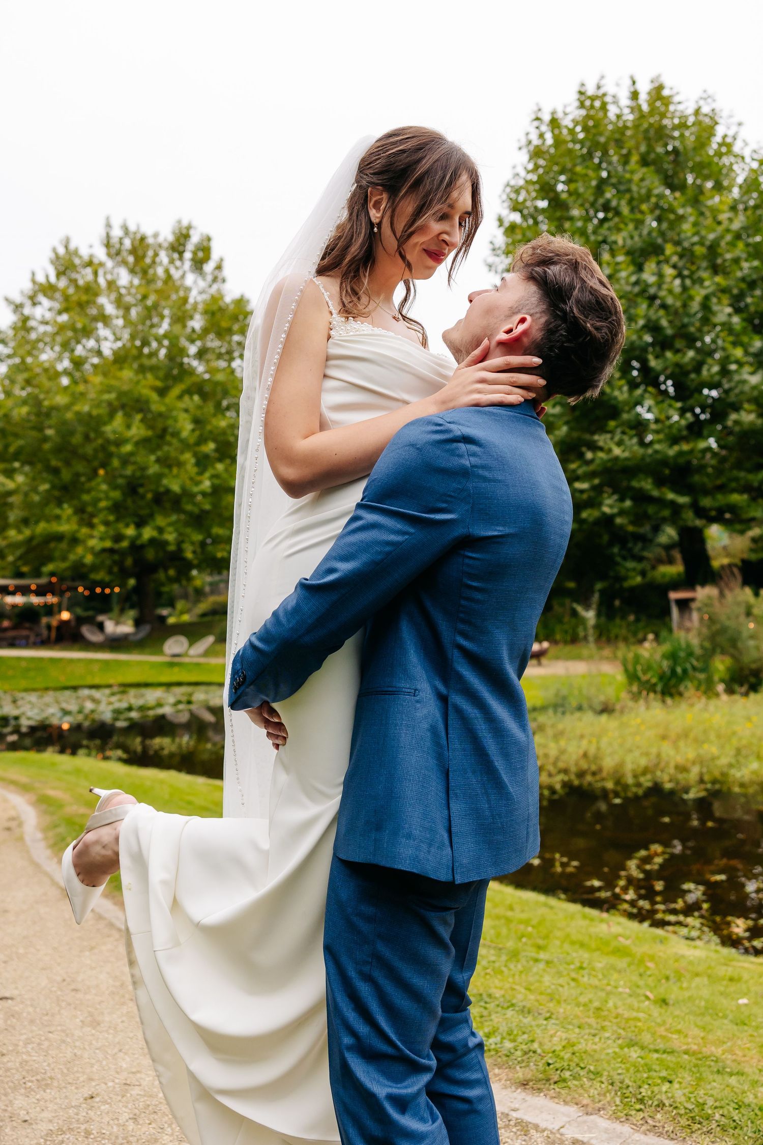 Groom lifting smiling bride in white wedding dress near a scenic garden pond.