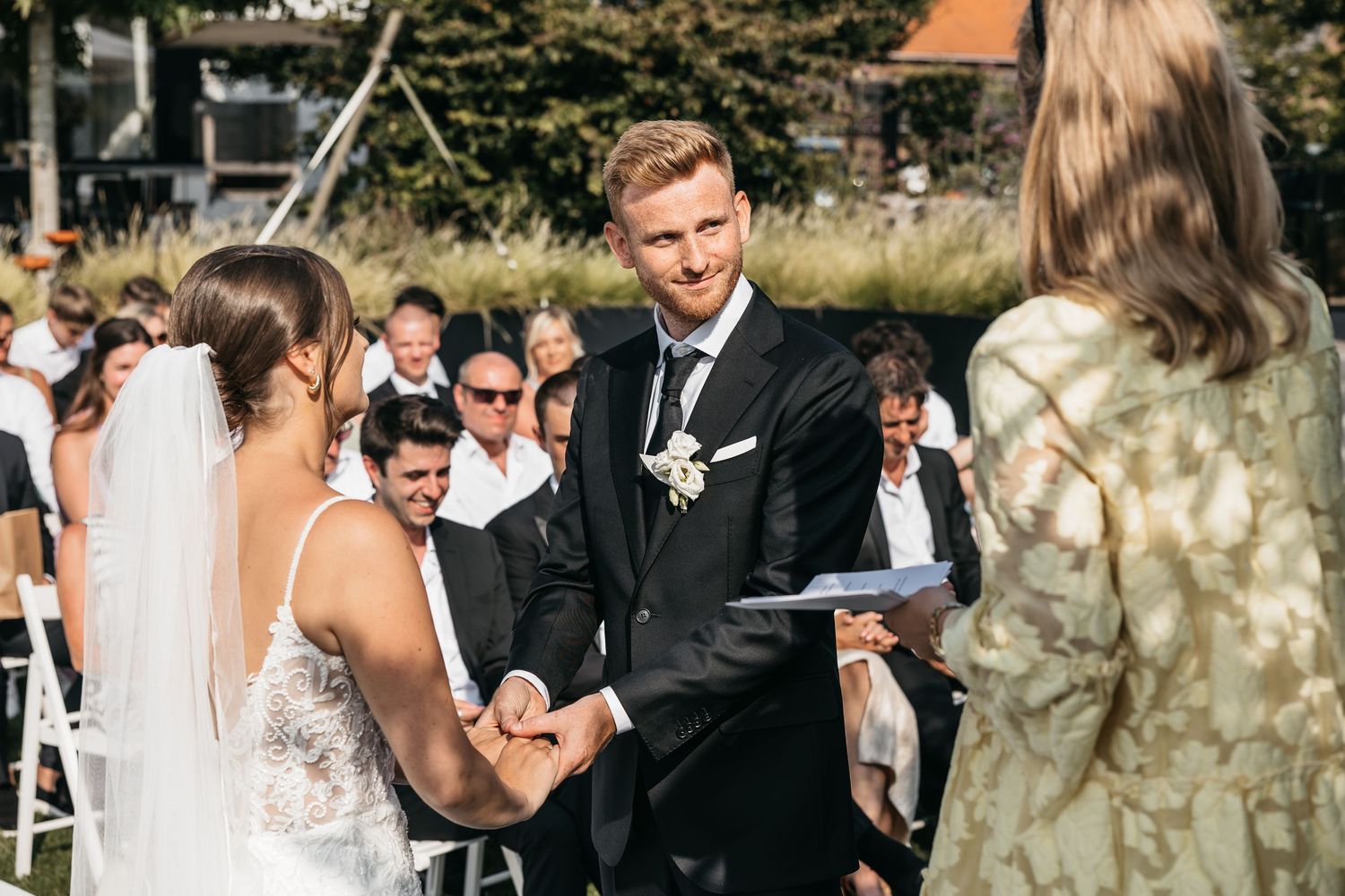 Groom smiles warmly while exchanging rings with bride at elegant outdoor wedding ceremony.