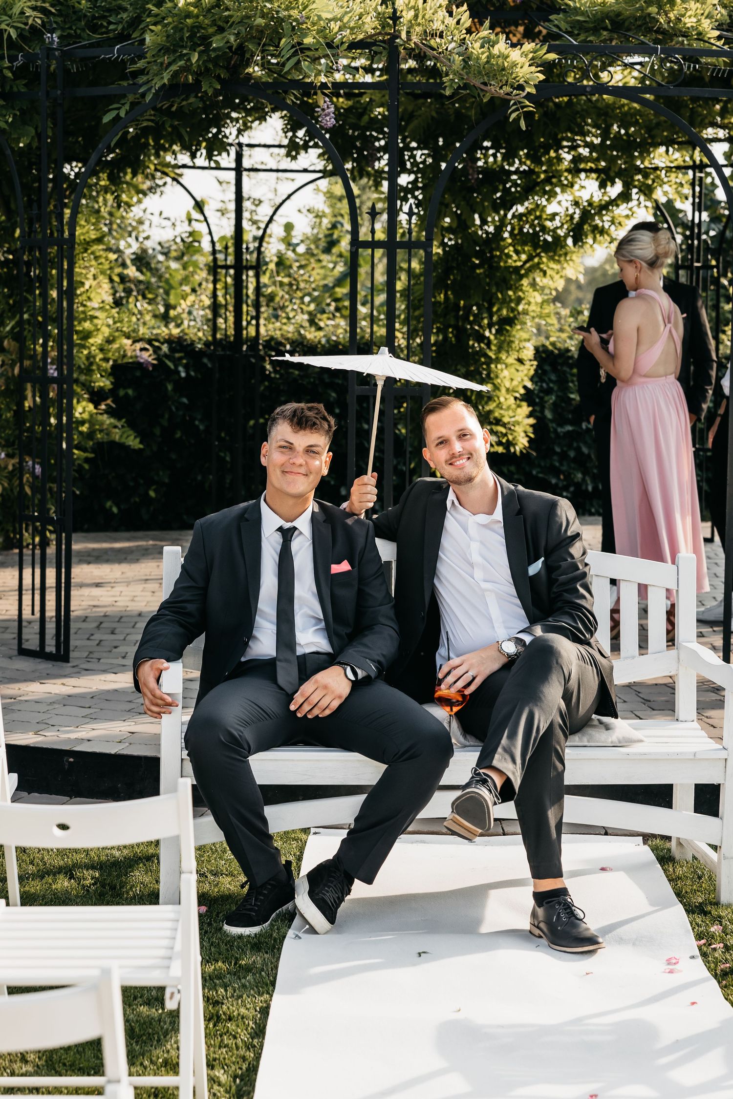Two young male wedding guests sit on white chairs holding a decorative umbrella outdoors.