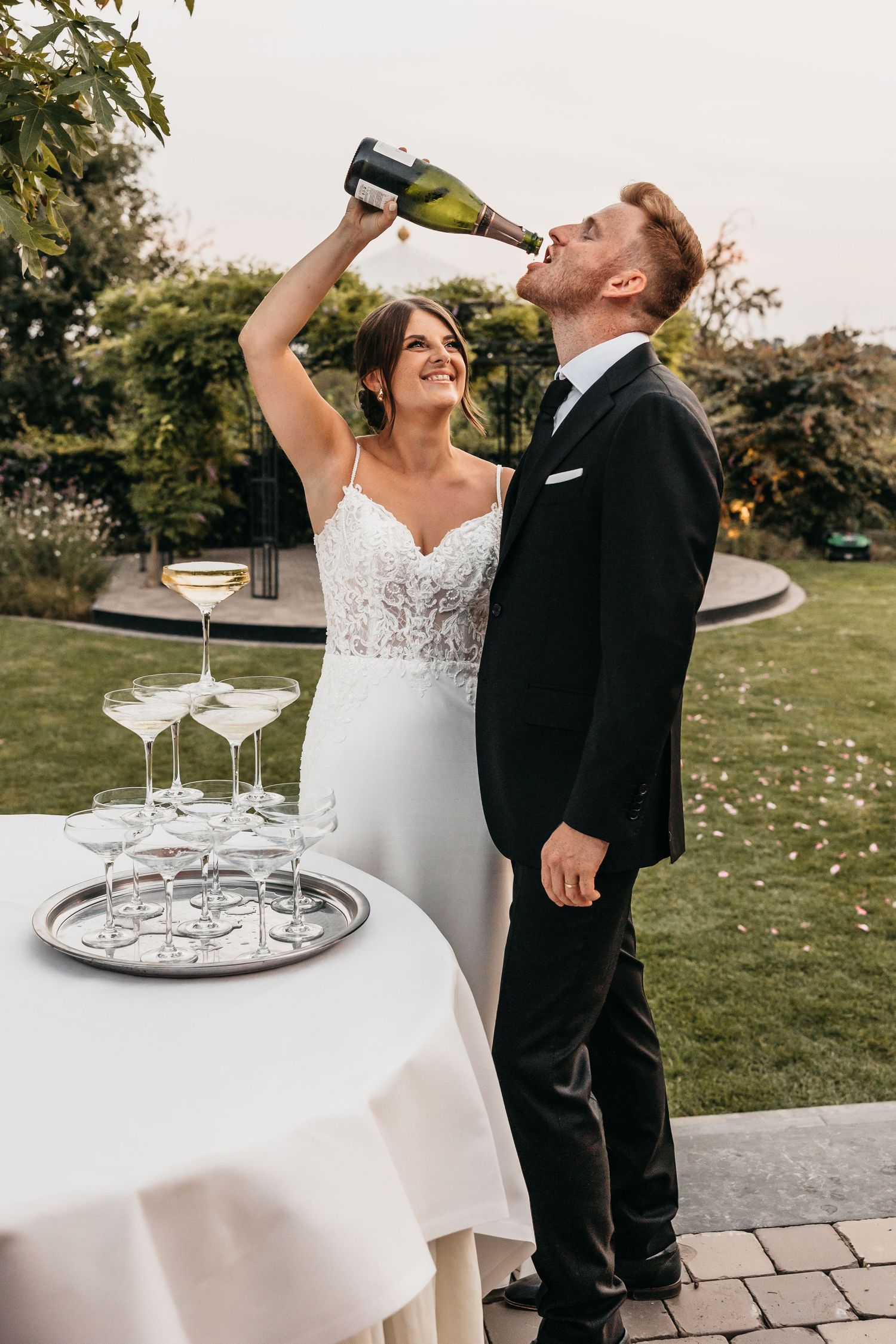 Playful bride pouring champagne bottle into grooms mouth at elegant outdoor wedding reception.